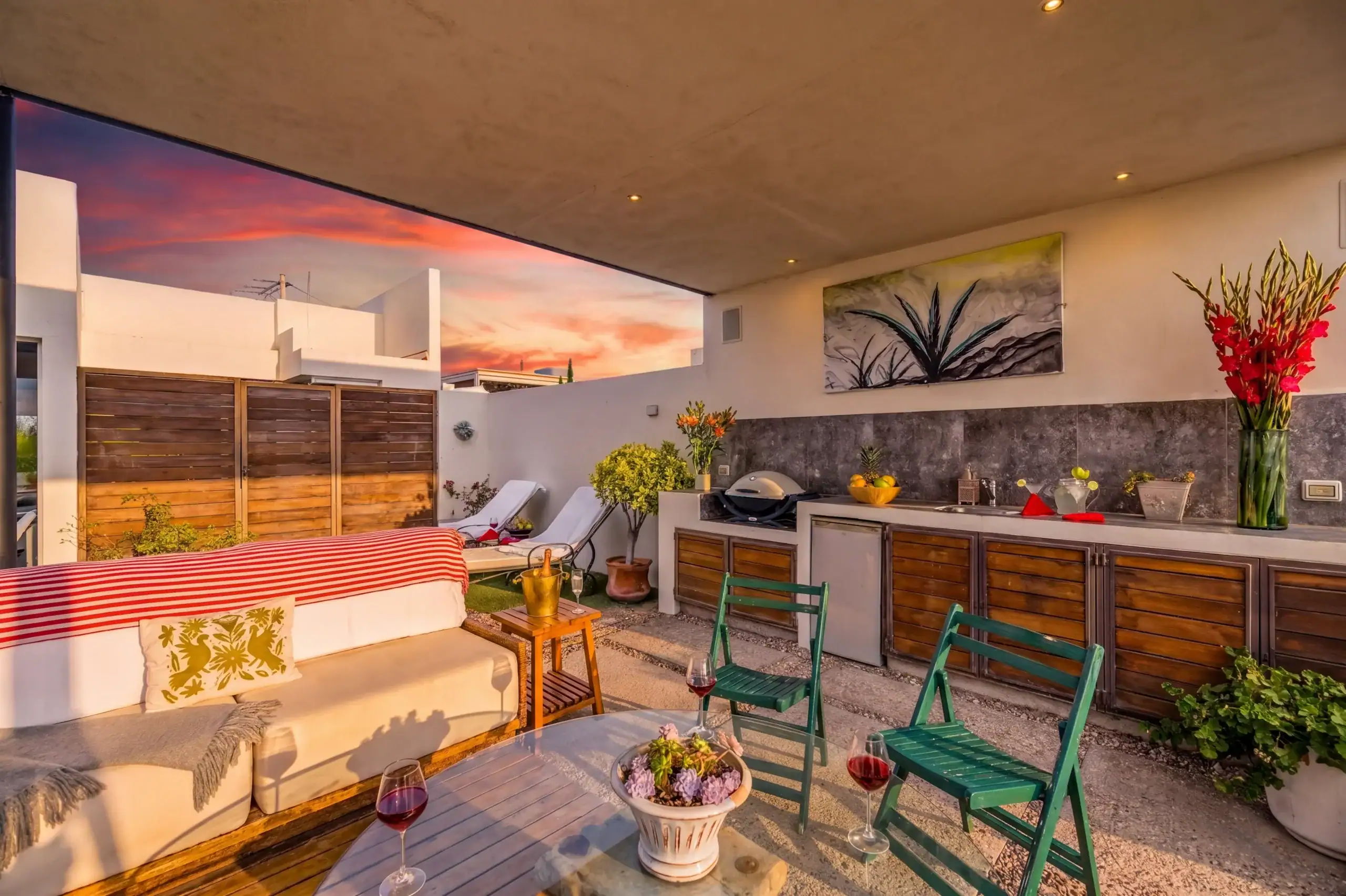 Outdoor patio at sunset with white seating, a red-striped throw, green metal chairs, wine glasses, and a mural on the wall, San Miguel de Allende