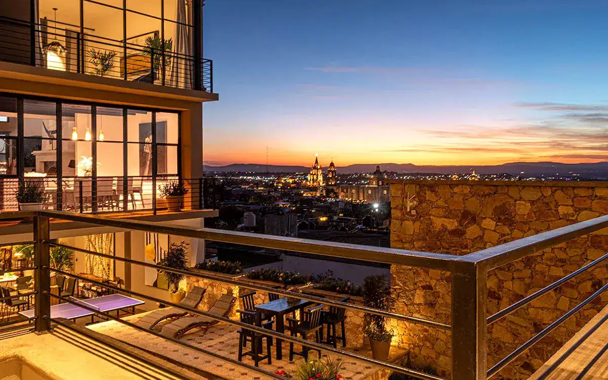 Modern rooftop terrace at dusk with glass walls, warm indoor lights, and a metal railing overlooking a city skyline and sunset.
