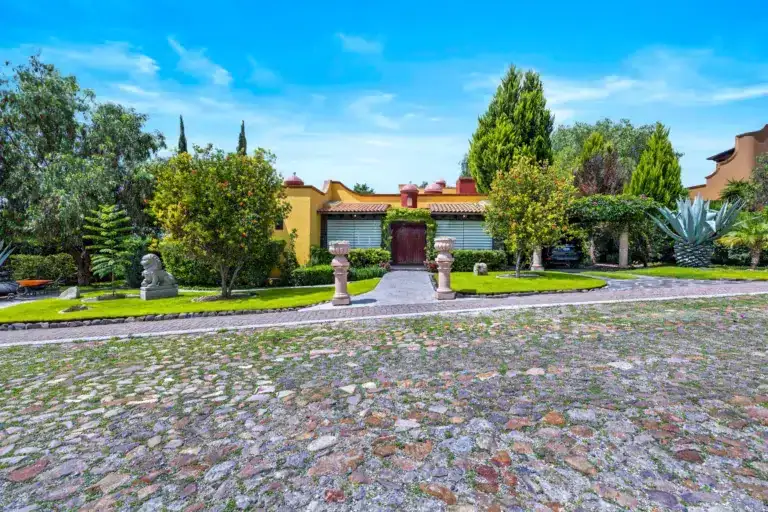 Front yard of a Mediterranean-style house with yellow walls, a red door, manicured hedges, and a cobblestone driveway under a bright blue sky, San Miguel de Allende
