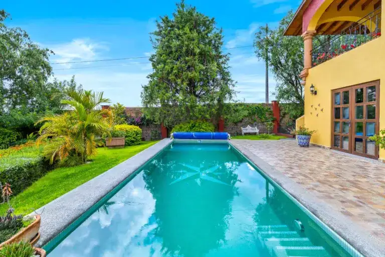 Rectangular pool with turquoise water bordered by stone, in a sunny backyard with a yellow house and lush plants nearby, San Miguel de Allende