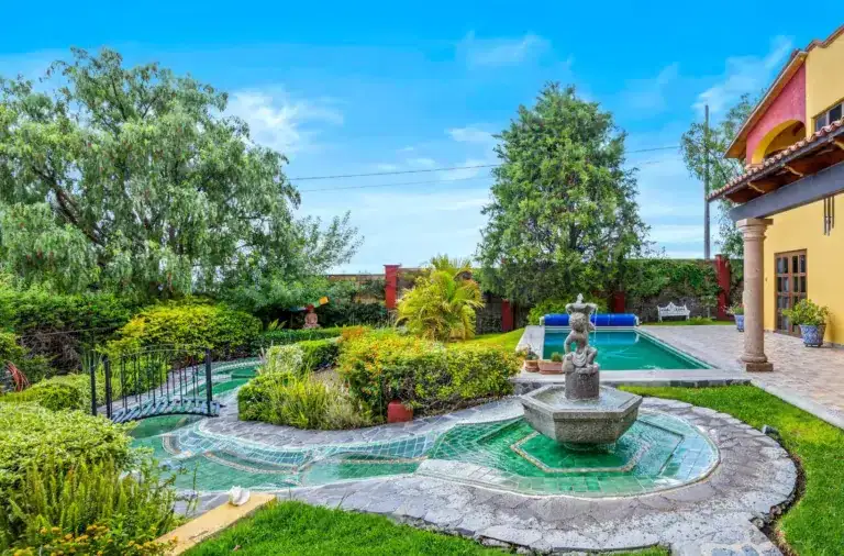 Sunny backyard with a rectangular pool, stone fountain, lush plants, and a yellow house with a columned patio on the right, San Miguel de Allende