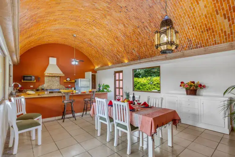 Open-concept kitchen and dining room with a brick vaulted ceiling, orange wall, and a hanging lantern; table set for a meal with red napkins and white chairs, San Miguel de Allende