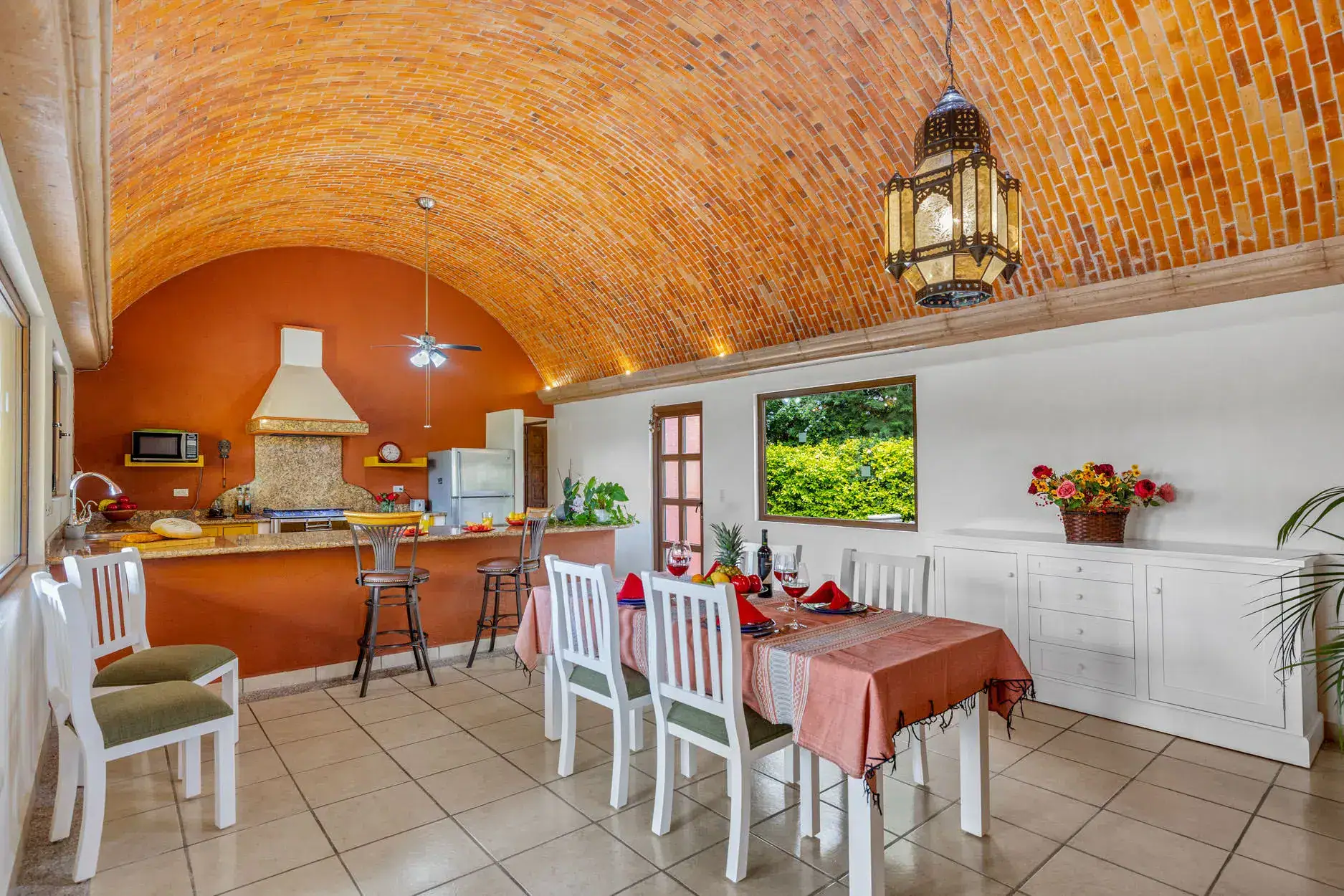 Open-concept kitchen and dining room with a brick vaulted ceiling, orange wall, and a hanging lantern; table set for a meal with red napkins and white chairs, San Miguel de Allende