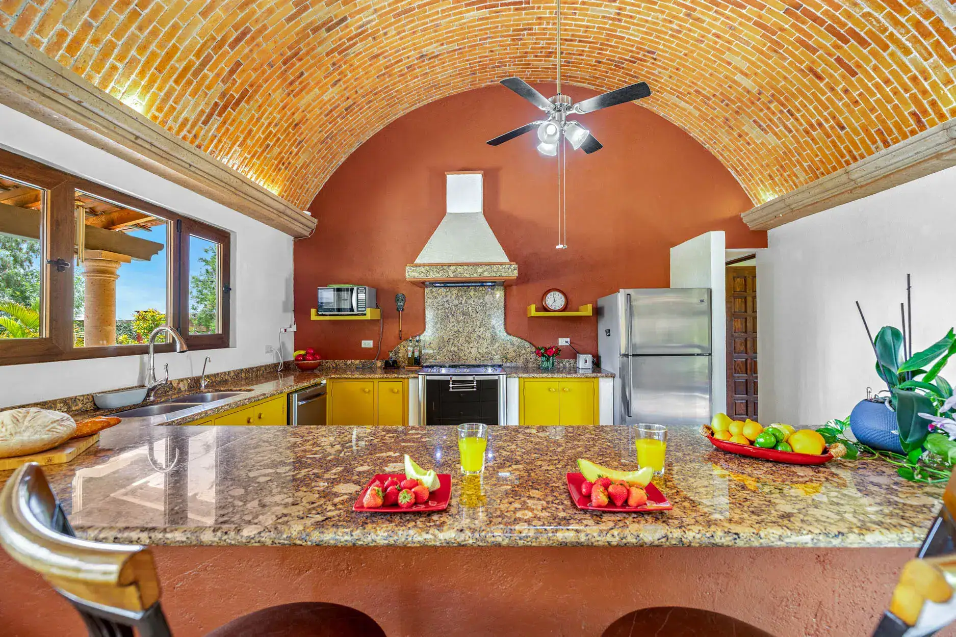 Bright kitchen with an arched brick ceiling, yellow lower cabinets, granite countertops, and stainless steel appliances; fruit on the island and windows on the left, San Miguel de Allende