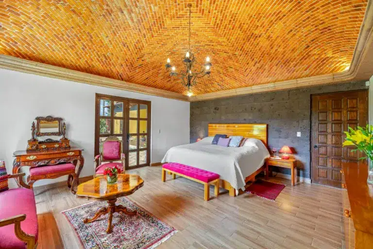 Luxury bedroom with a brick vaulted ceiling and ornate chandelier above a wooden-framed bed with white linens and pink cushions, San Miguel de Allende