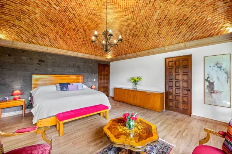 Bedroom with a vaulted brick ceiling, ornate chandelier, and a large bed with gray bedding and colorful throw pillows, wood panel headboard visible on left side, San Miguel de Allende