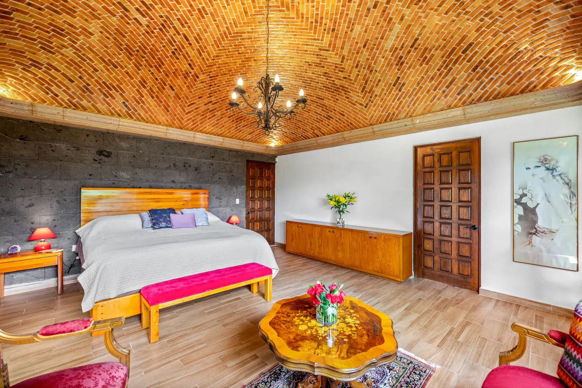 Bedroom with a vaulted brick ceiling, ornate chandelier, and a large bed with gray bedding and colorful throw pillows, wood panel headboard visible on left side, San Miguel de Allende
