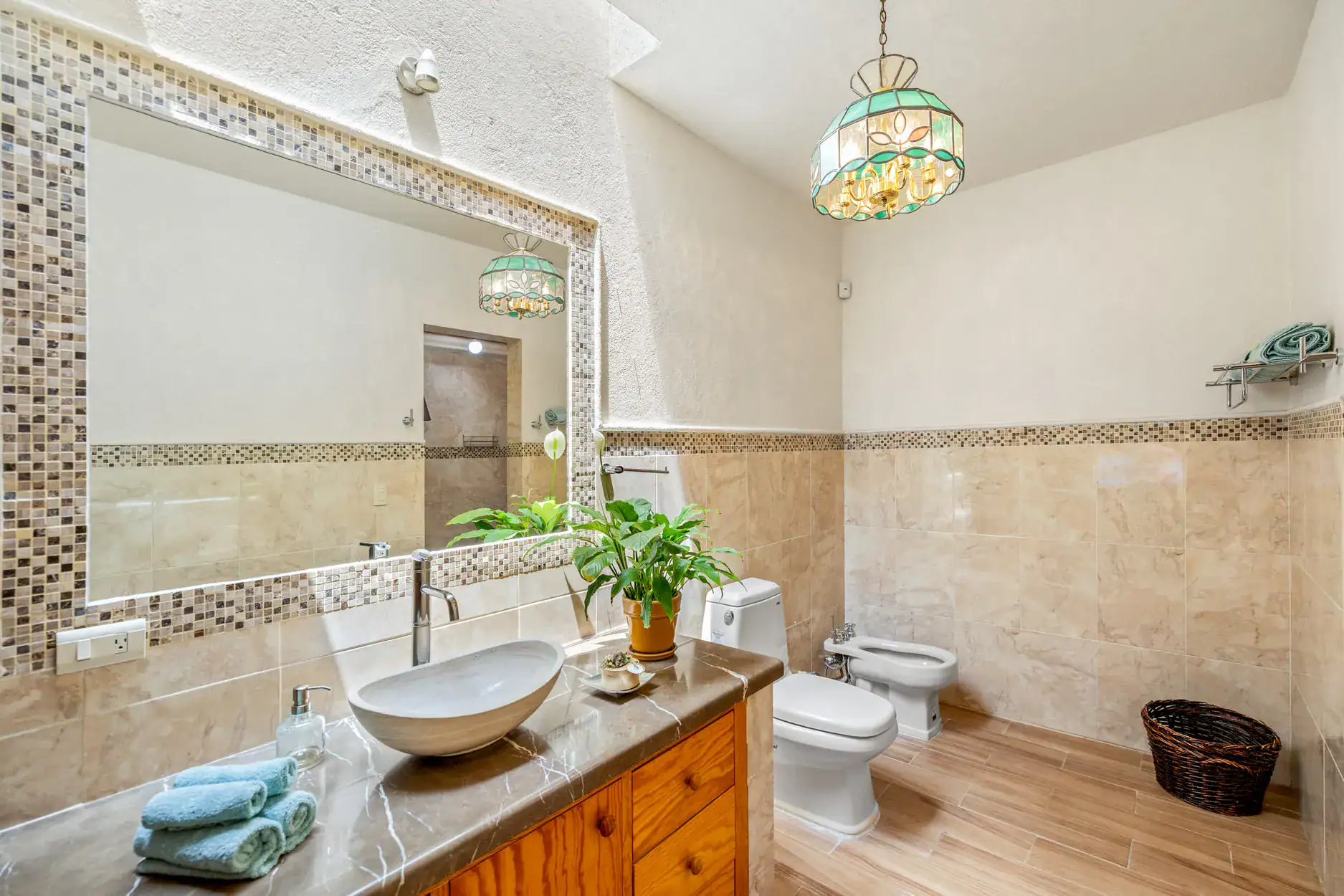 Bathroom with marble countertop, double round sinks, wooden vanity, mosaic tile trim, and a large mirror with a potted plant nearby, San Miguel de Allende