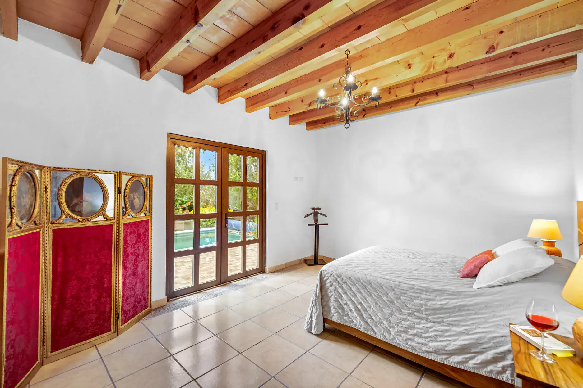 Bright bedroom with exposed wooden ceiling beams, glass French doors to a garden, and a red ornate folding screen beside the door, San Miguel de Allende