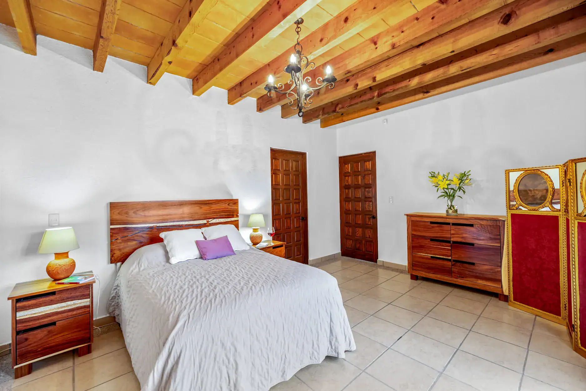 Bedroom with a beamed wooden ceiling, chandelier, and a large bed with white bedding and purple pillows, San Miguel de Allende