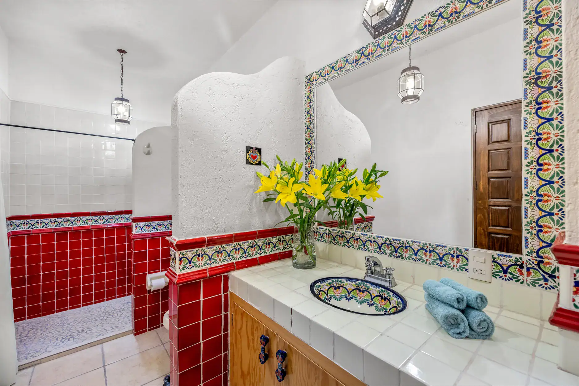 Bright bathroom with red tiled walls and white counters; yellow lilies in a glass vase sit on the sink counter under a decorative mirror and colorful tile trim, San Miguel de Allende