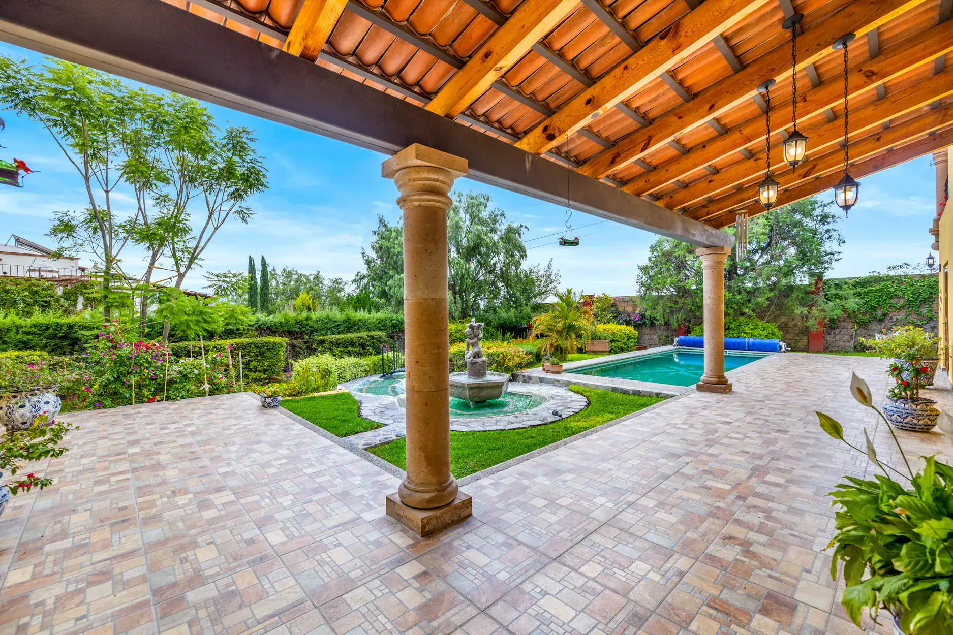 Covered patio with stone columns, hanging lanterns, and a garden view including a fountain and pool in the background, San Miguel de Allende