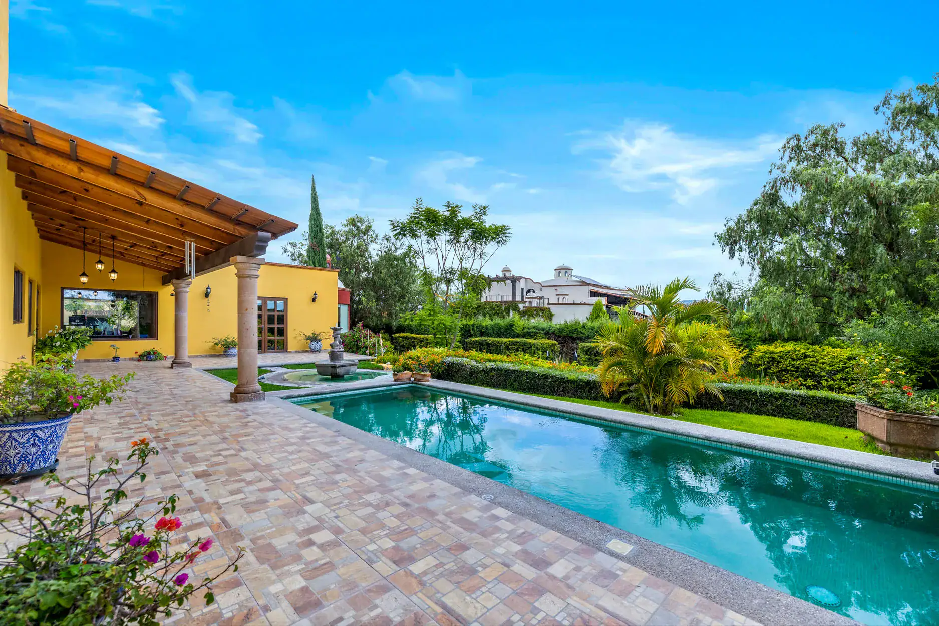 Yellow villa with a wooden pergola, stone columns, and a long rectangular pool surrounded by a tiled patio and potted plants, San Miguel de Allende