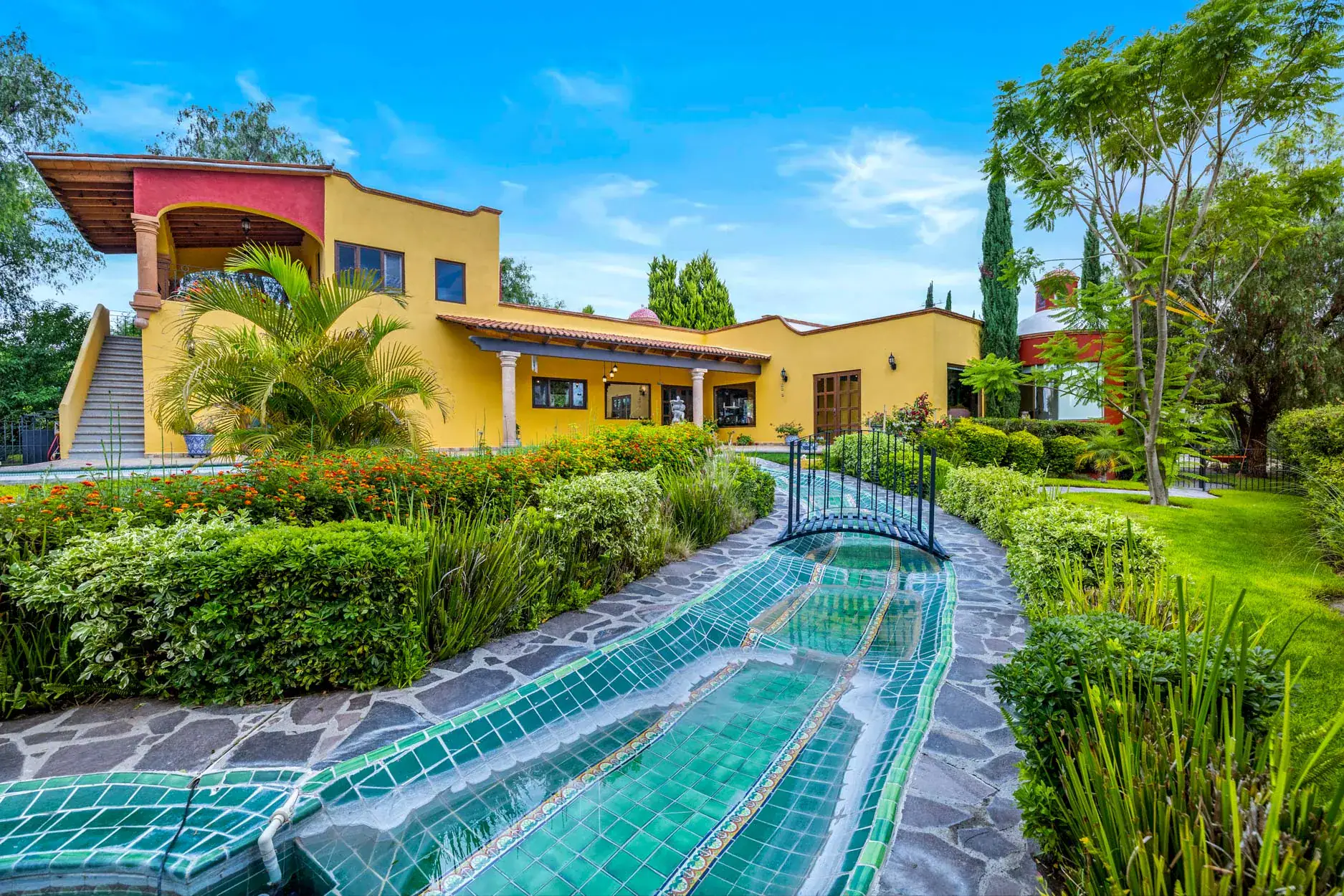 Mediterranean-style yellow villa with tiled canal and lush garden under a bright blue sky, San Miguel de Allende