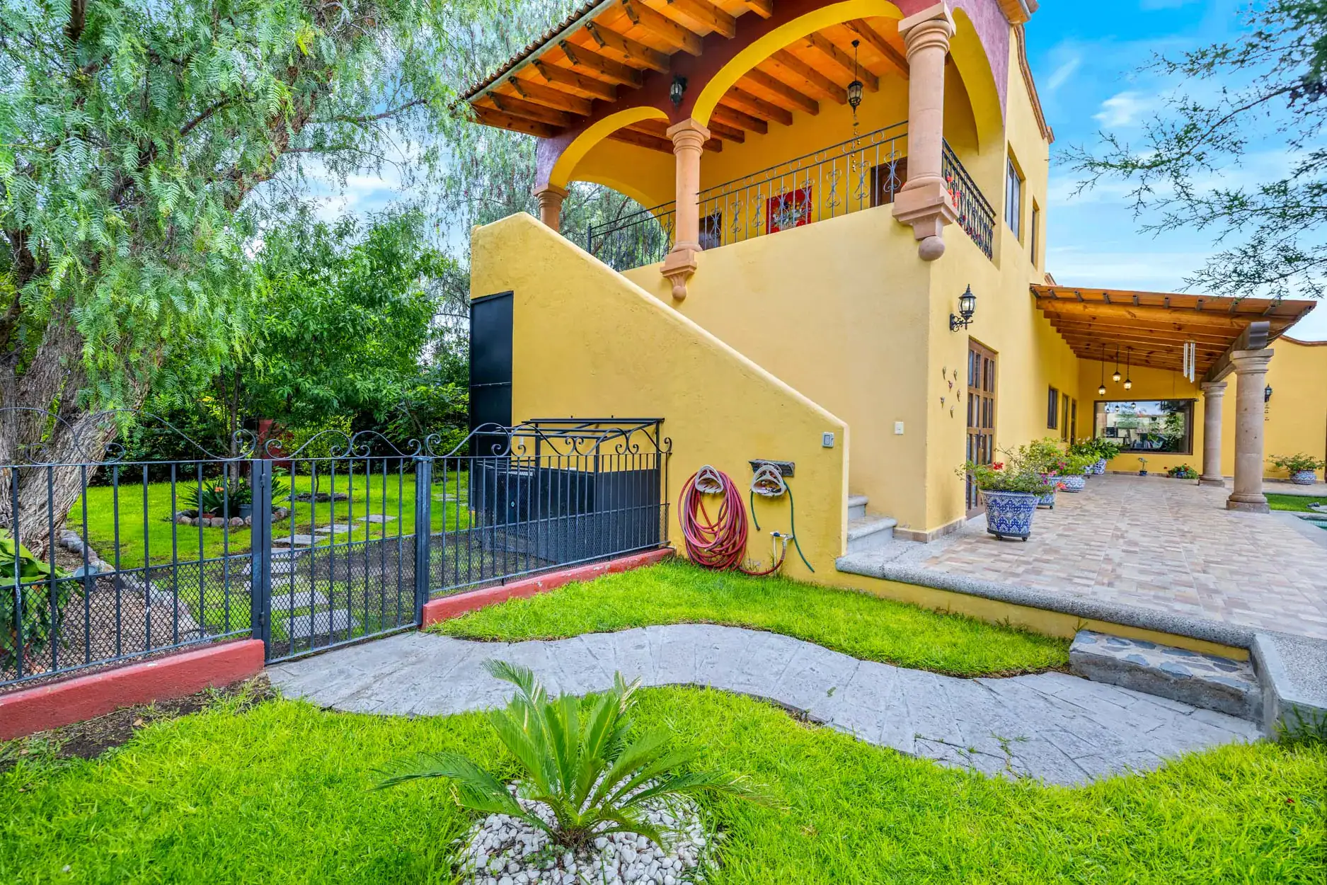Yellow two-story house with arched porch and wrought-iron balcony, wooden beams, and a tiled patio surrounded by green lawn and trees; garden hoses hang on the wall, San Miguel de Allende