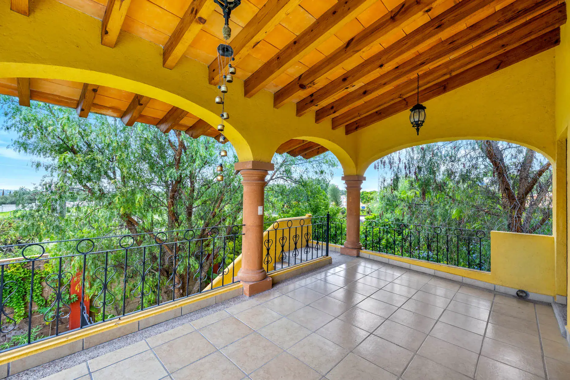 Yellow arched veranda with wooden beams, hanging bells, and a lantern, overlooking green trees and a garden outside, San Miguel de Allende