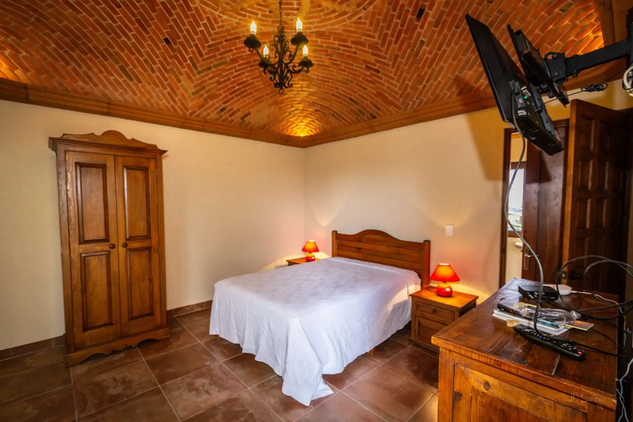 Cozy bedroom with a brick vaulted ceiling, chandelier, wooden wardrobe, bed with white bedspread, and red bedside lamps, San Miguel de Allende