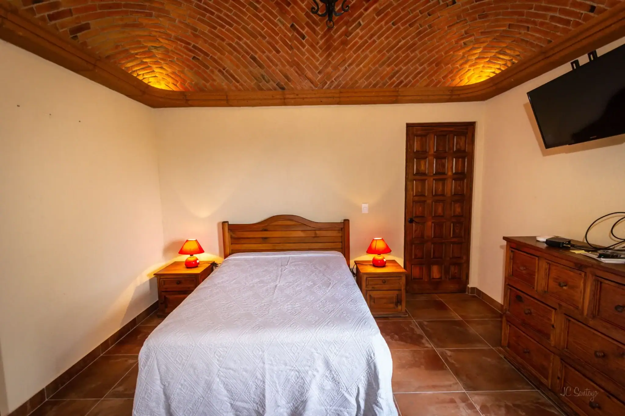 Wooden bedroom with a brick vaulted ceiling and warm amber lighting; a bed flanked by two matching nightstands with red lamps, a wooden door on the wall, and a dresser with a mounted TV to the right, San Miguel de Allende