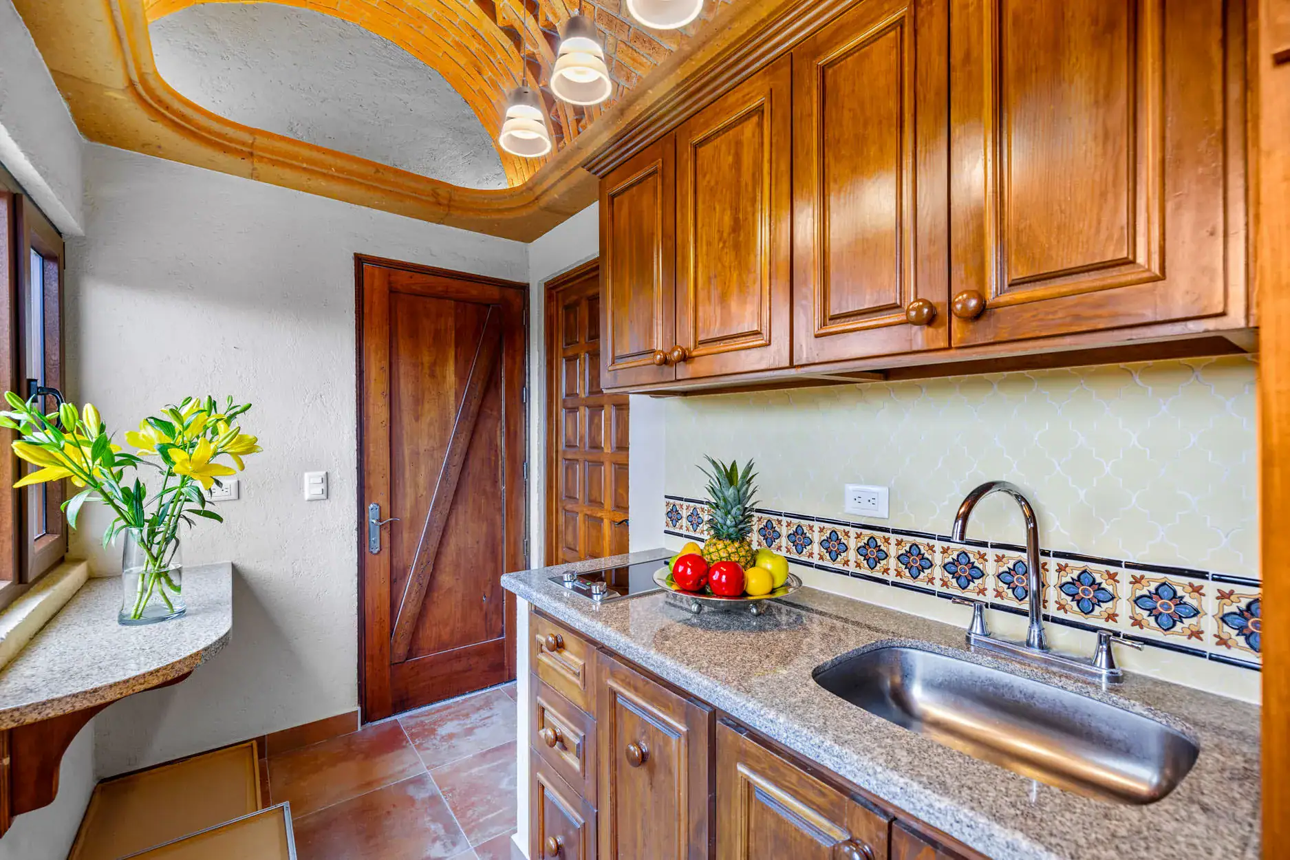 Rustic kitchen with dark wood cabinets, granite countertop, and a fruit bowl near the sink under patterned tile backsplash art, San Miguel de Allende