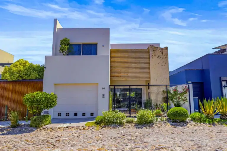 Modern two-story white house with a tall vertical wall, wooden slat screen, and a black gate under a bright blue sky, San Miguel de Allende
