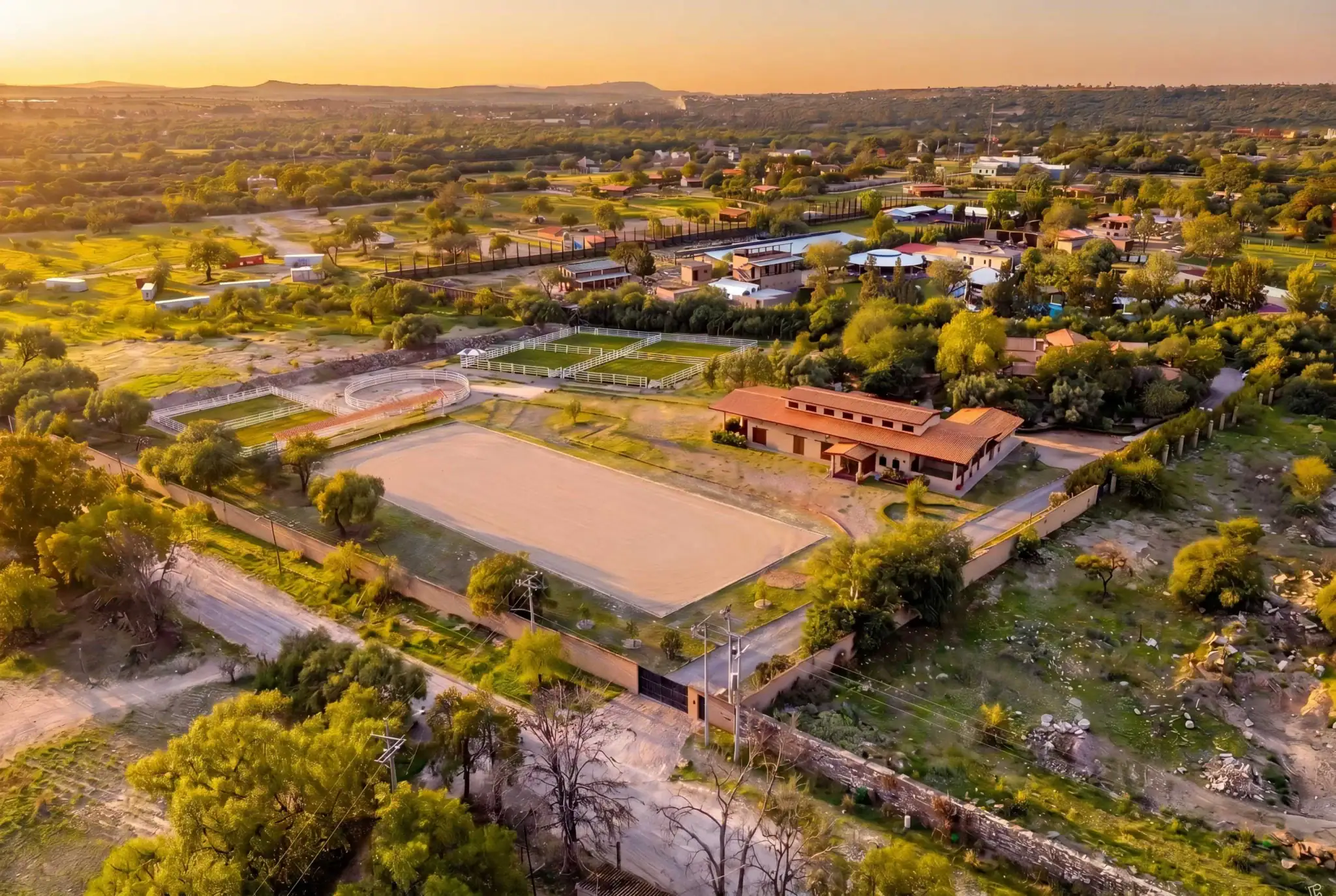 Aerial view of a small town at sunset, with rodeo arenas, fences, and scattered buildings amid green trees and fields, San Miguel de Allende
