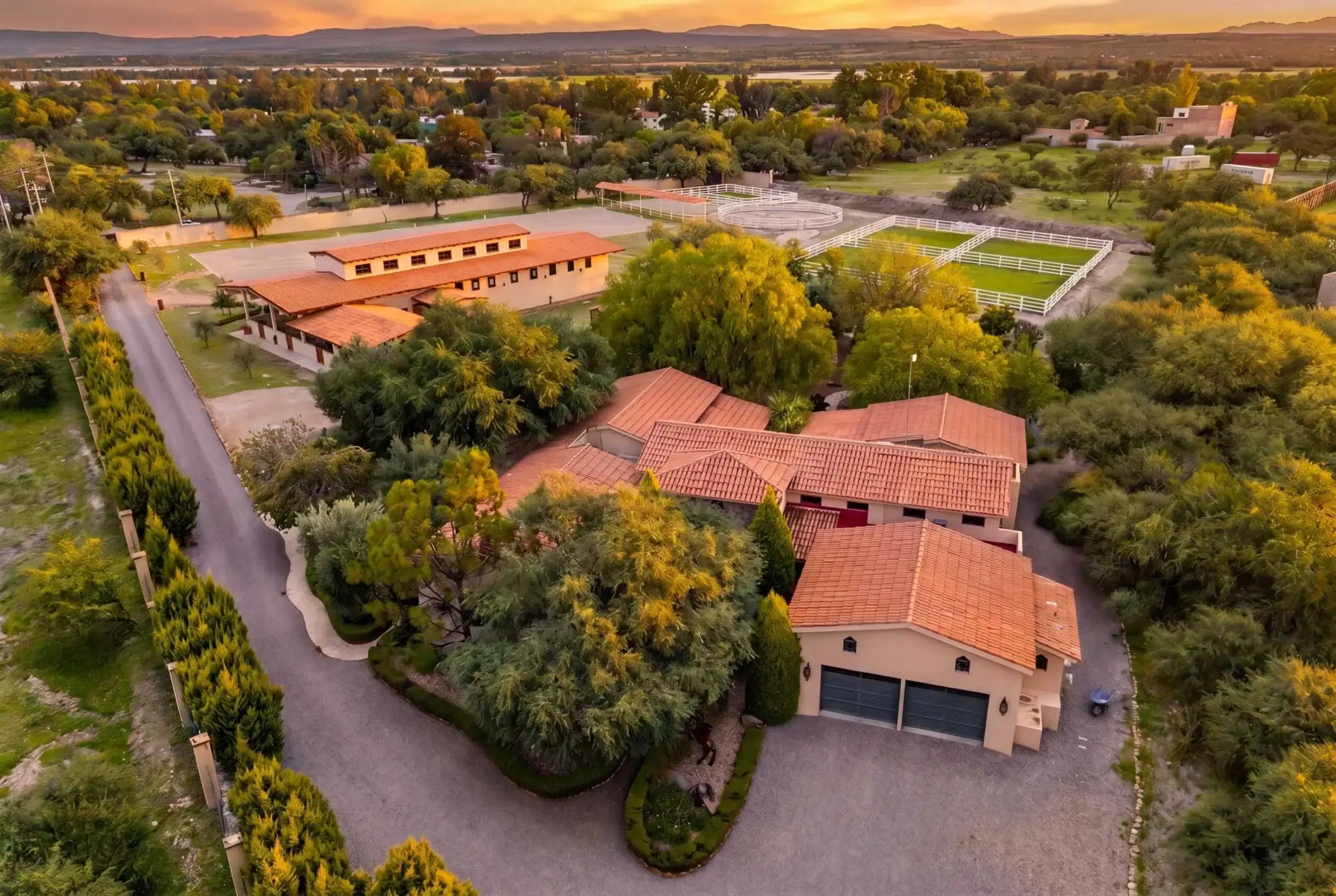 Aerial view of a suburban ranch area with terracotta roof buildings, white fencing, riding arenas, and sunset lighting, San Miguel de Allende