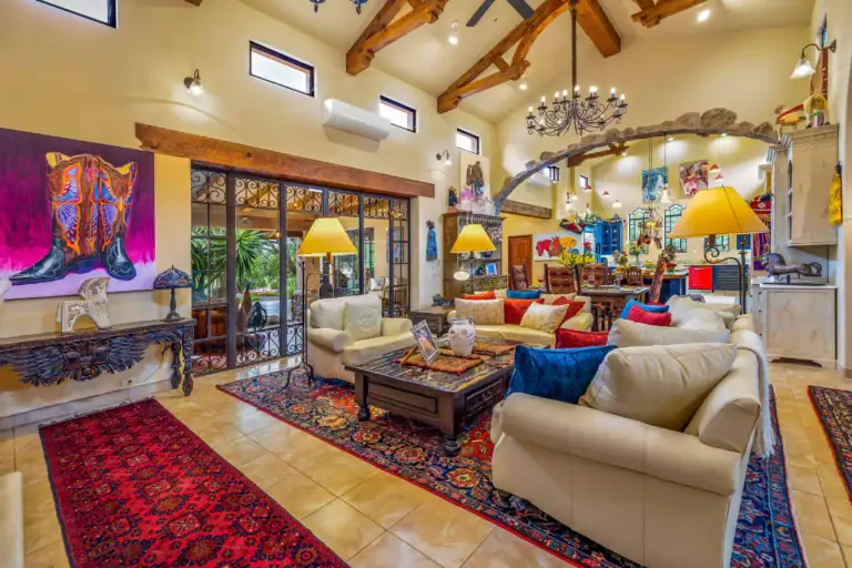 Spacious living room with cream sofas, colorful pillows, and red patterned rugs under a wooden coffee table; exposed wooden beams and a wrought-iron chandelier overhead, San Miguel de Allende