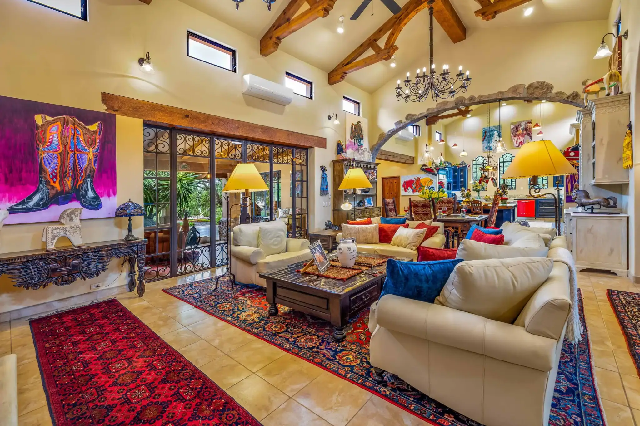 Spacious living room with cream sofas, colorful pillows, and red patterned rugs under a wooden coffee table; exposed wooden beams and a wrought-iron chandelier overhead, San Miguel de Allende