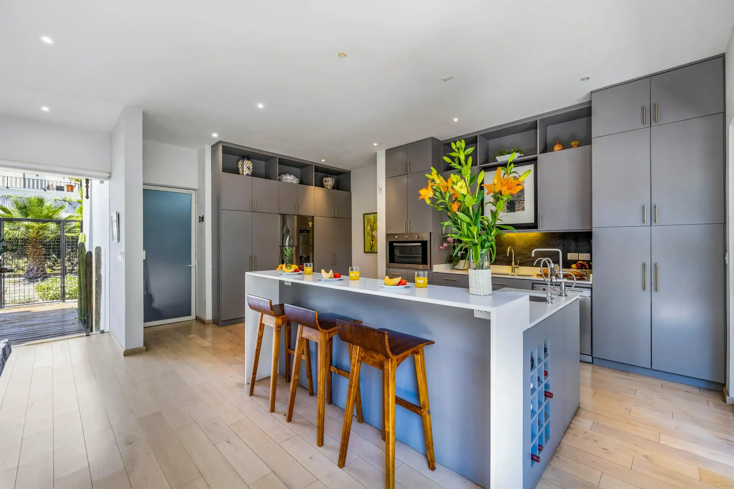 Modern kitchen with a white island and gray cabinetry, bar stools, and a large vase with orange flowers as a centerpiece; built-in appliances and open shelves line the back wall, with a sliding door to the outdoors visible to the left, San Miguel de Allende