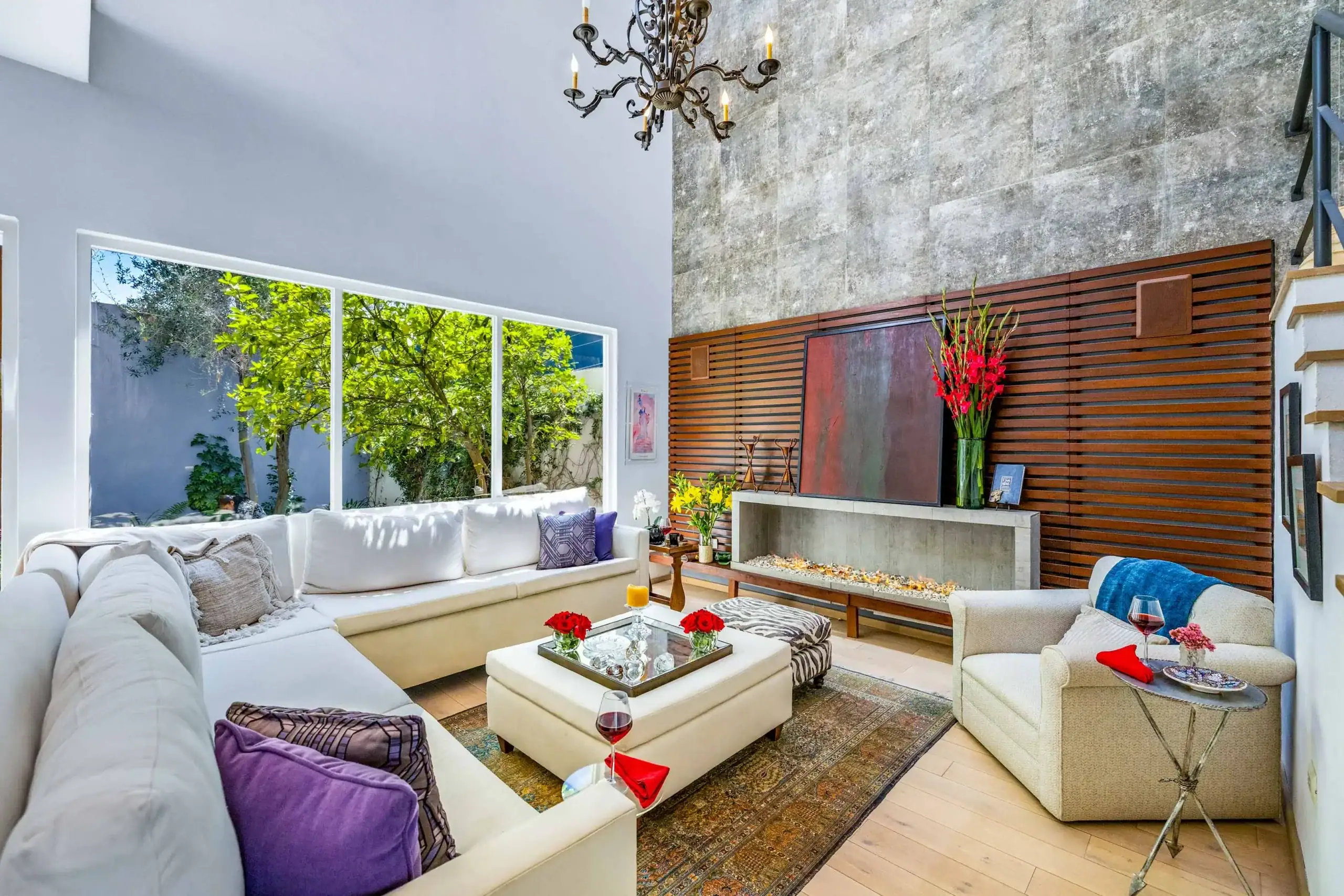 Bright modern living room with white sectional sofa, purple pillows, and a glass coffee table holding red flowers and wine glasses, near a large windowed wall with greenery outside, San Miguel de Allende