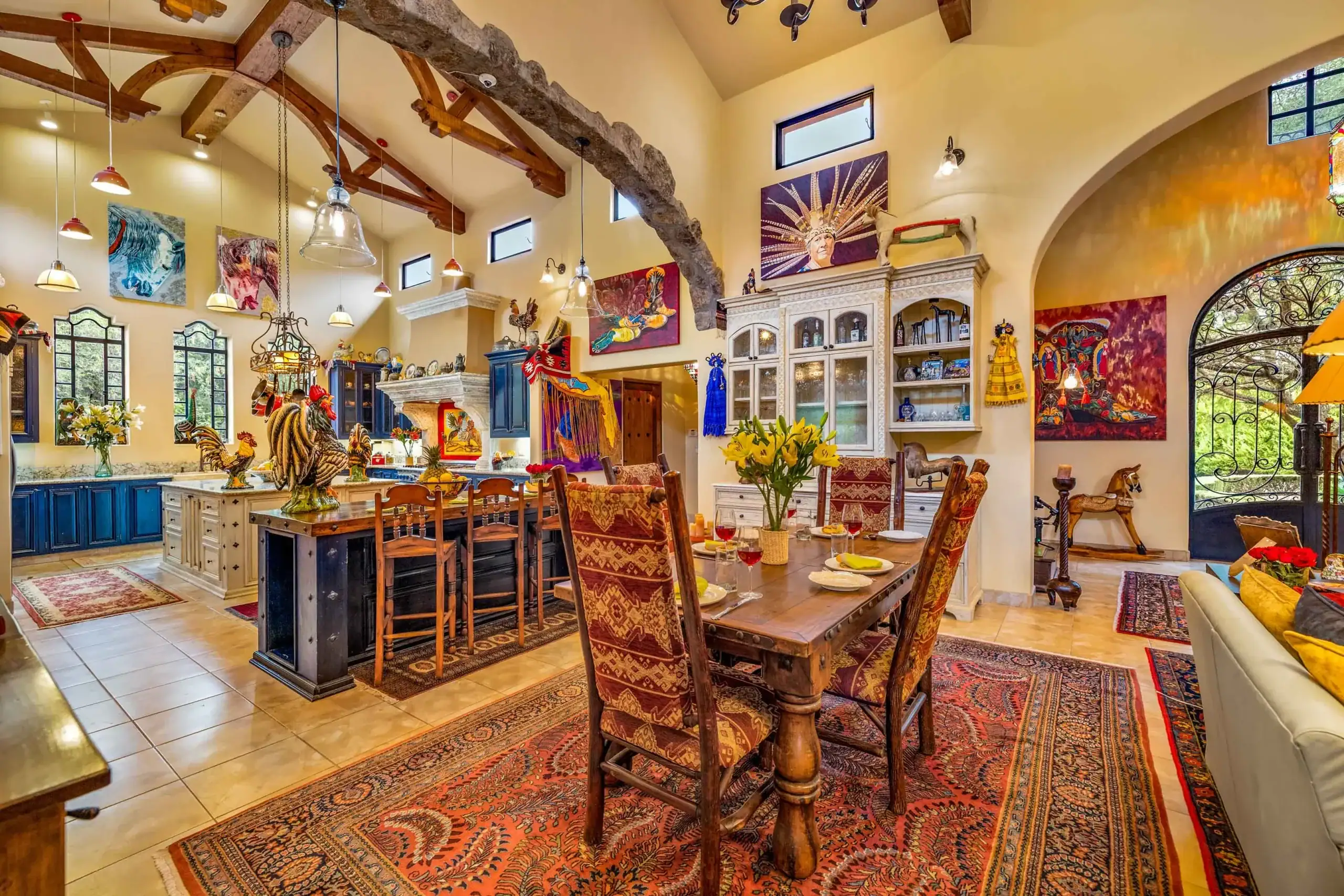 Eclectic kitchen-dining room with exposed wooden beams, warm yellow walls, blue cabinets, and colorful artwork and chandeliers, San Miguel de Allende