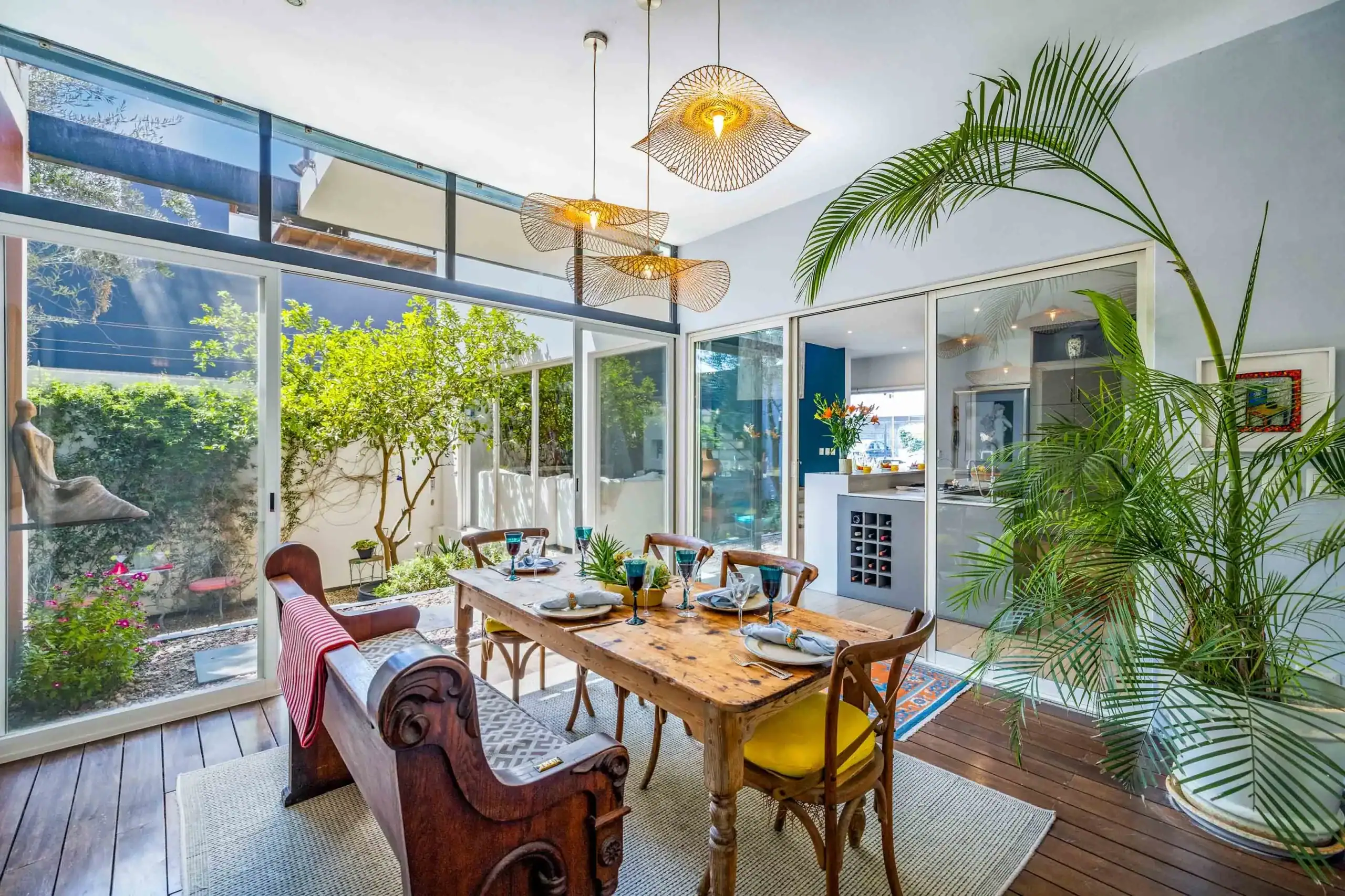 Bright dining room with a rustic wooden table set for eight, surrounded by carved chairs, glass walls, and tropical plants, San Miguel de Allende