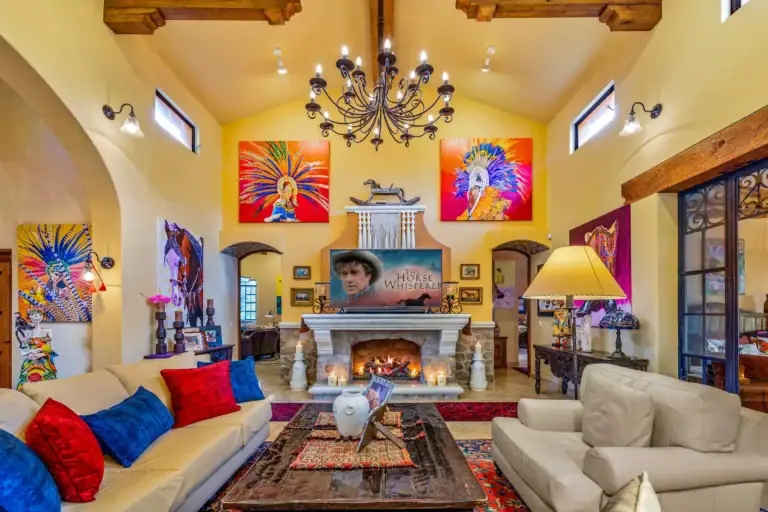 Bright living room with a wrought-iron chandelier, yellow walls, and colorful pop-art paintings above a stone fireplace, flanked by beige sofas with red and blue cushions, San Miguel de Allende