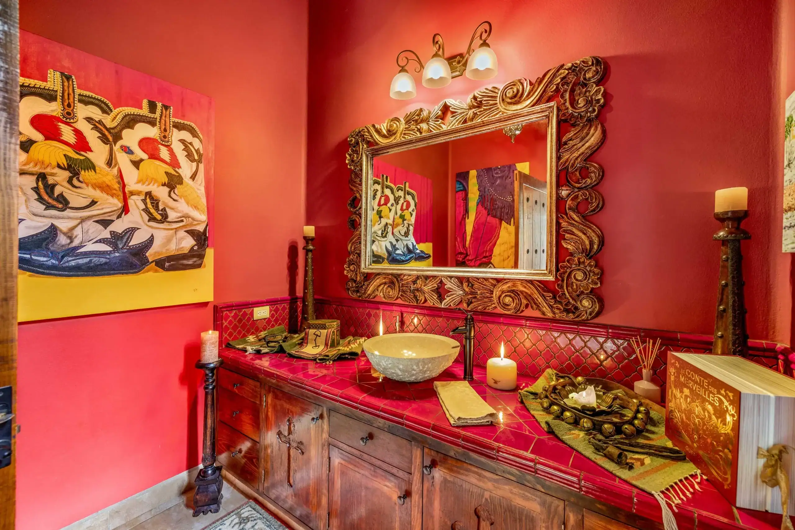 Red-painted dressing room with a large ornate carved-wood mirror above a red tiled counter, candles, and decorative objects, San Miguel de Allende