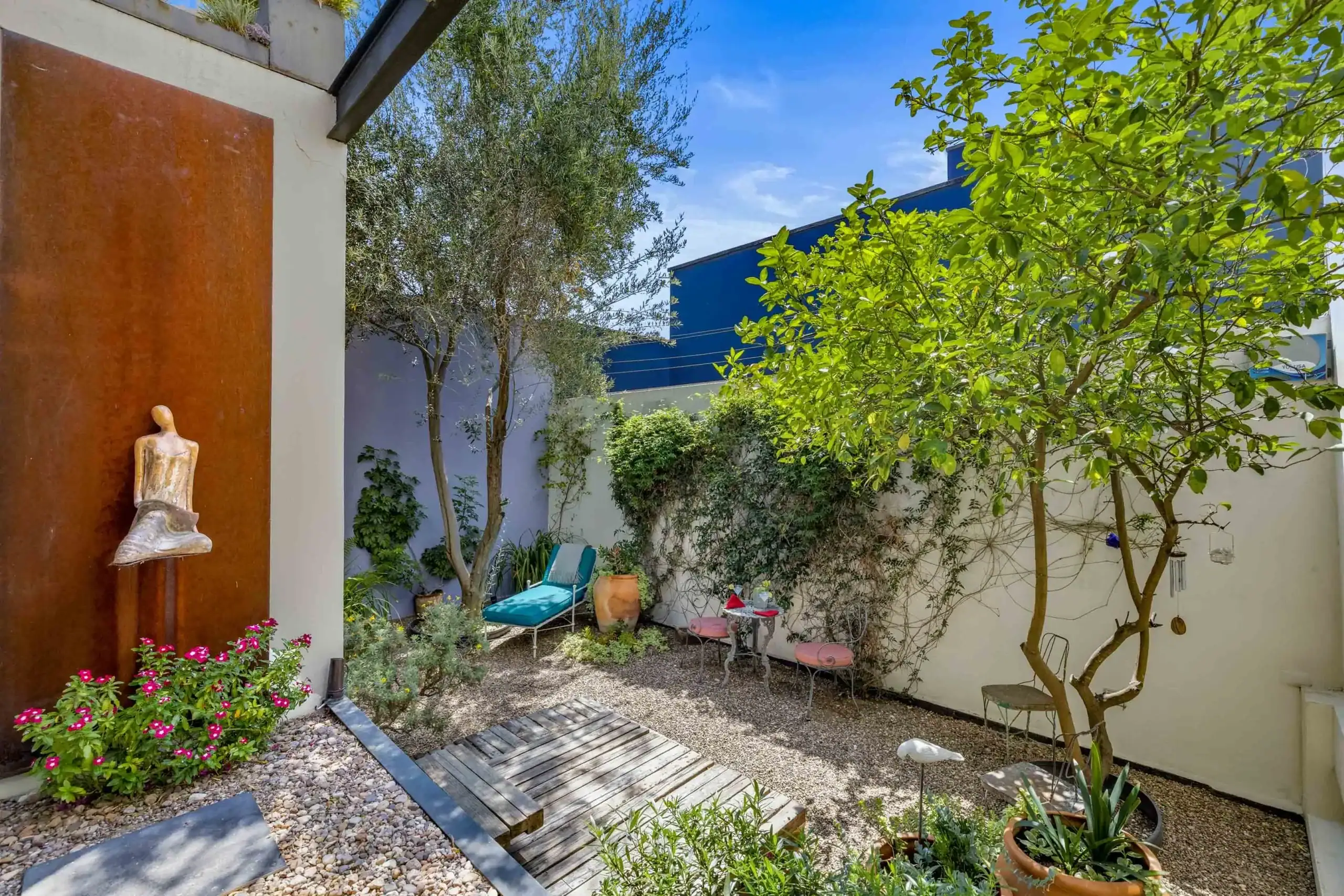 Sunny backyard courtyard with a wooden boardwalk, gravel ground, and a seating area among potted plants and trees, San Miguel de Allende