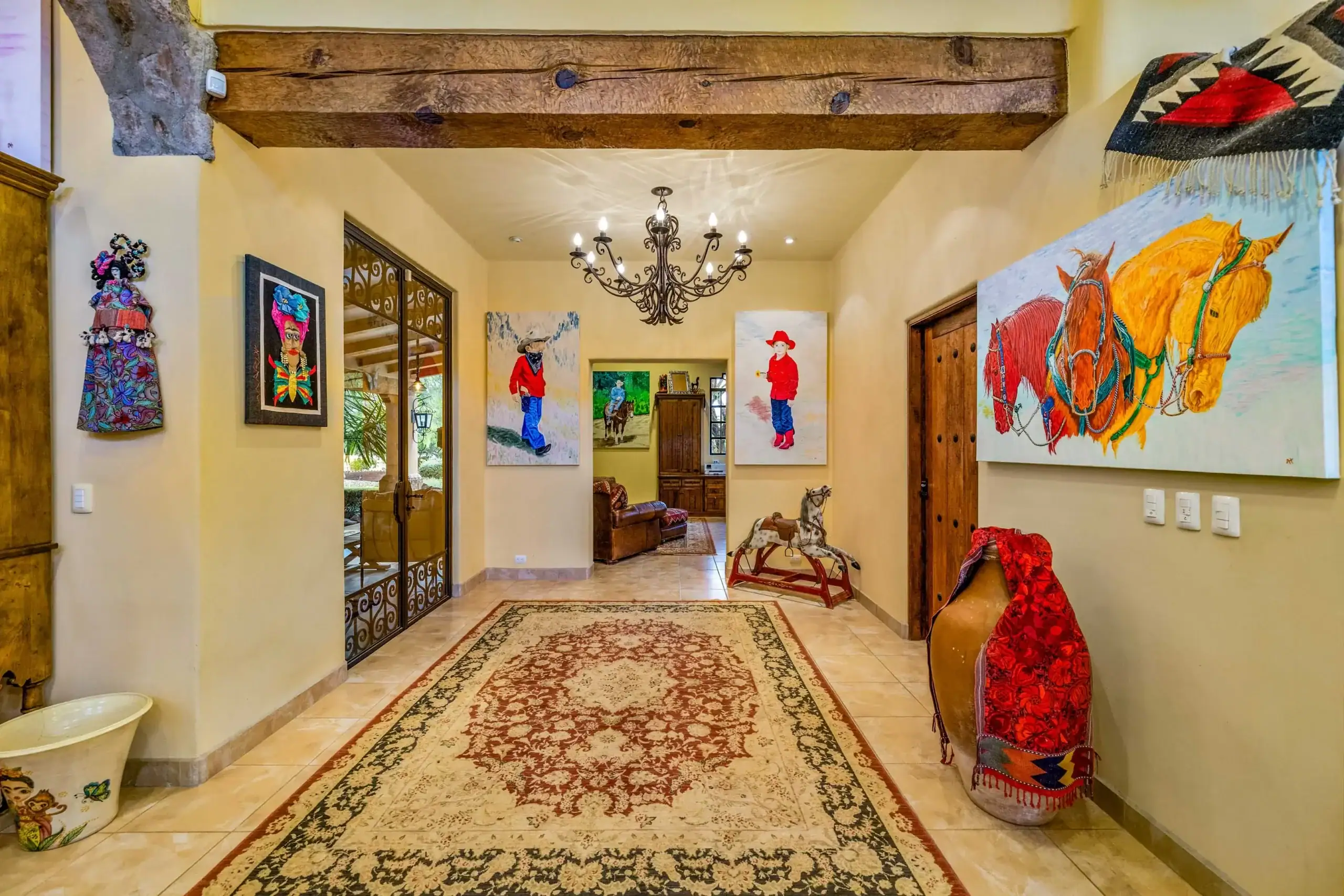 Bright foyer hallway with beige walls, a dark wood beam, a wrought-iron chandelier, and bold horse-themed paintings along the walls, San Miguel de Allende
