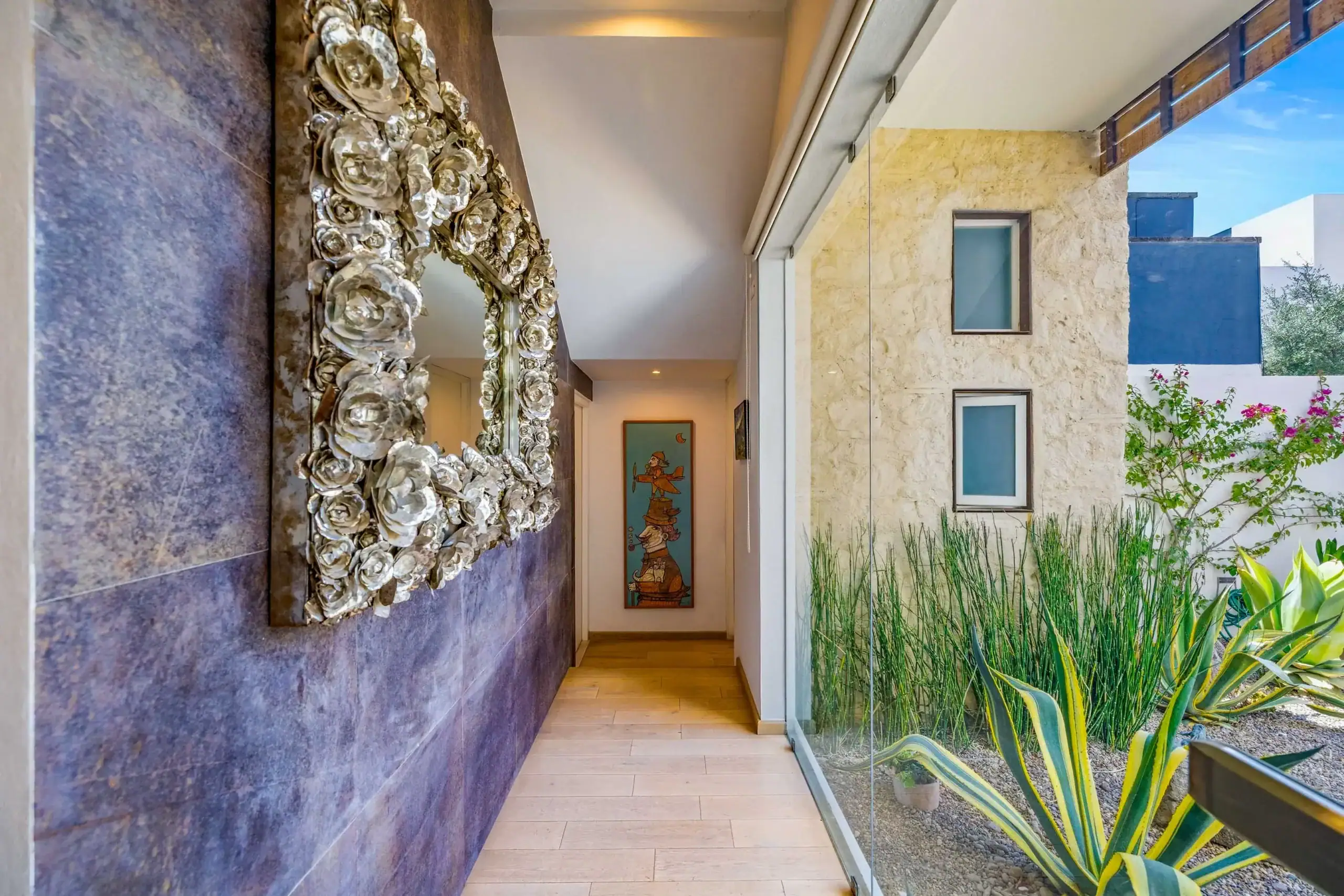 Indoor hallway featuring a large circular mirror frame of silver roses mounted on a dark stone wall; glass wall to the right looks into a courtyard with tall green plants, San Miguel de Allende