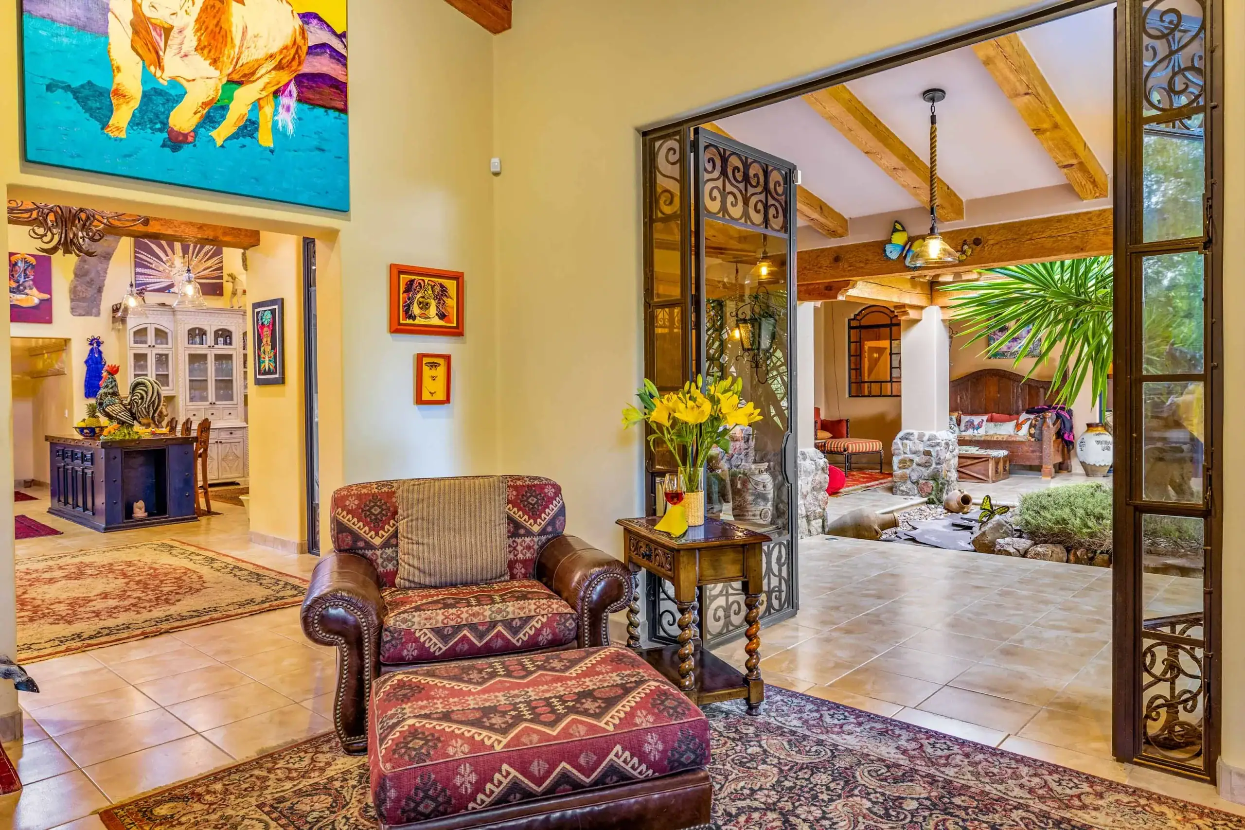 Cozy living room with patterned red armchairs, wooden side tables, and a tiled floor, opening to a sunlit patio with exposed wooden beams and greenery nearby, San Miguel de Allende