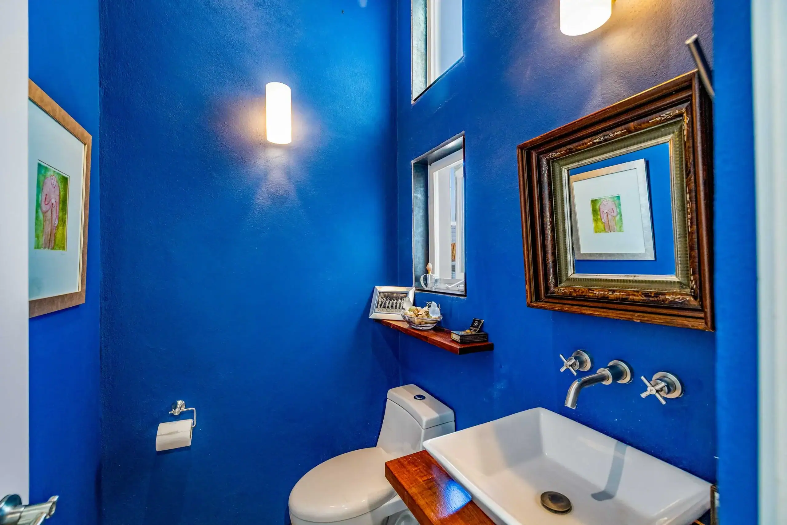 Blue bathroom with a white rectangular sink on a wooden countertop, a white toilet, and chrome fixtures under framed art, San Miguel de Allende