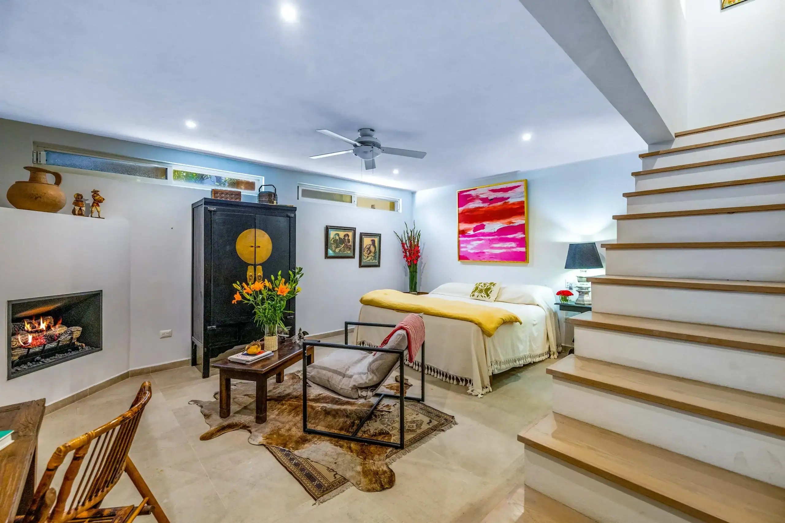 Living area with a bed, black cabinet, fireplace, and a bright pink abstract painting; staircase on the right adds height to the room, San Miguel de Allende