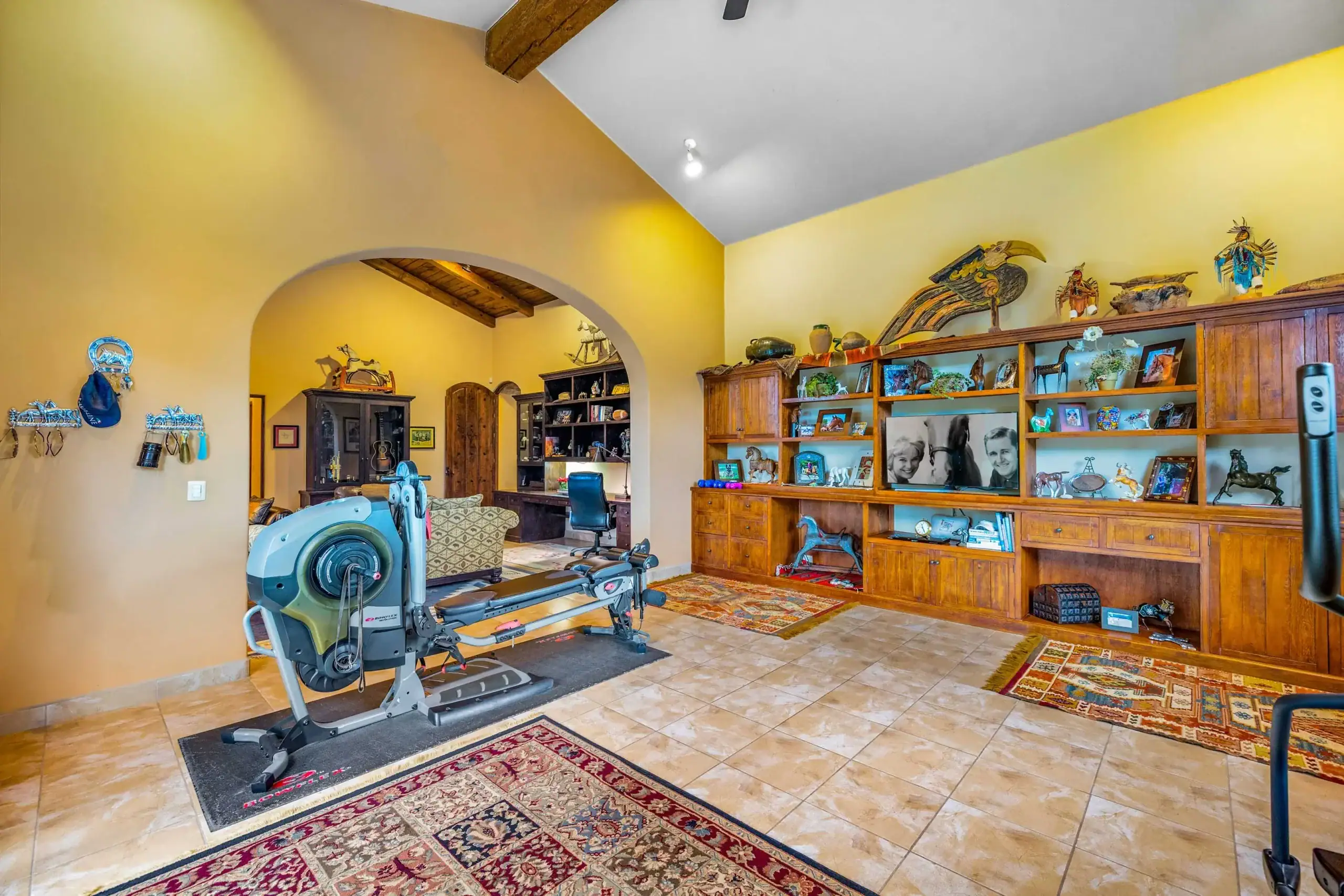 Home gym in a warm yellow living room with a cardio machine, arched doorway, and a tall wooden display cabinet filled with decor, San Miguel de Allende