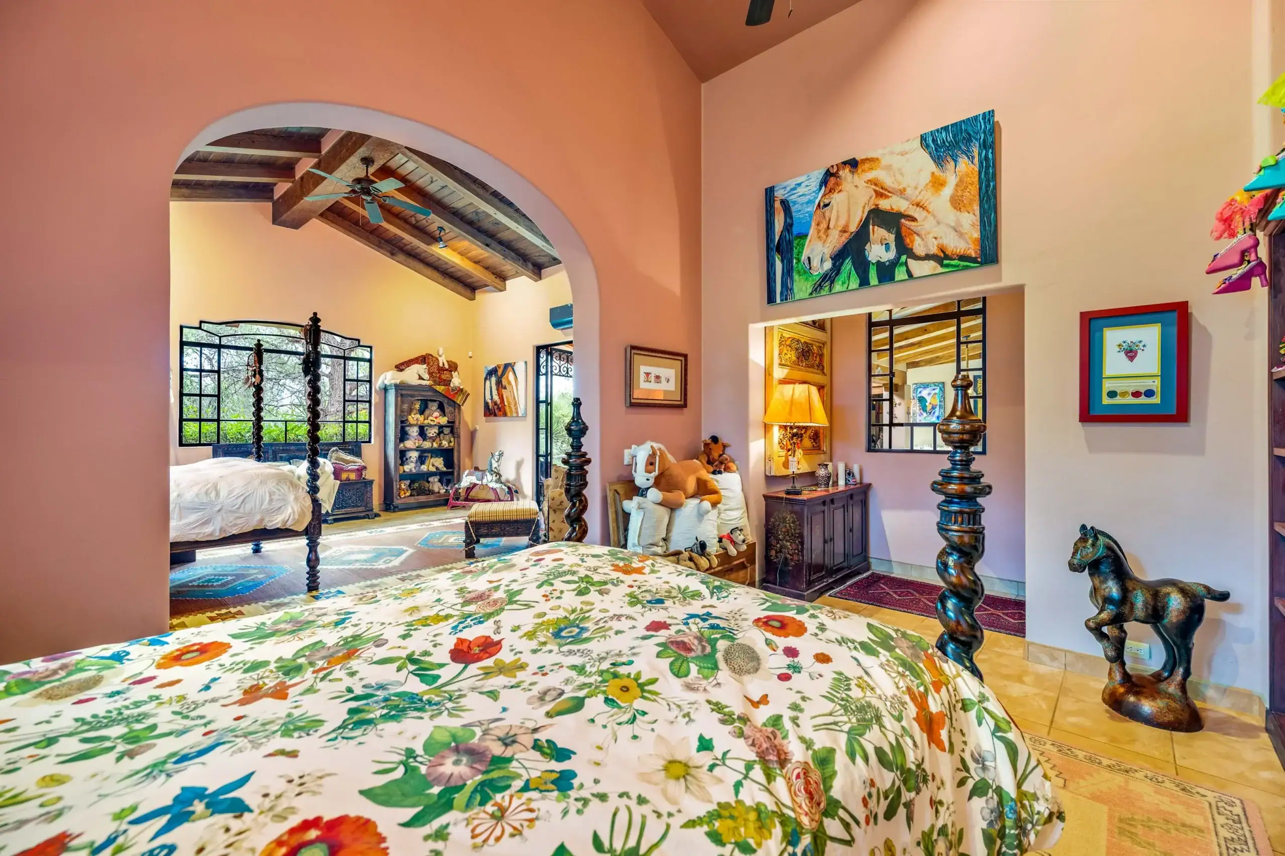 Colorful bedroom with floral bedspread, arched doorway, and dark wooden four-poster bed; stuffed animals and bright wall art create a playful, eclectic space, San Miguel de Allende