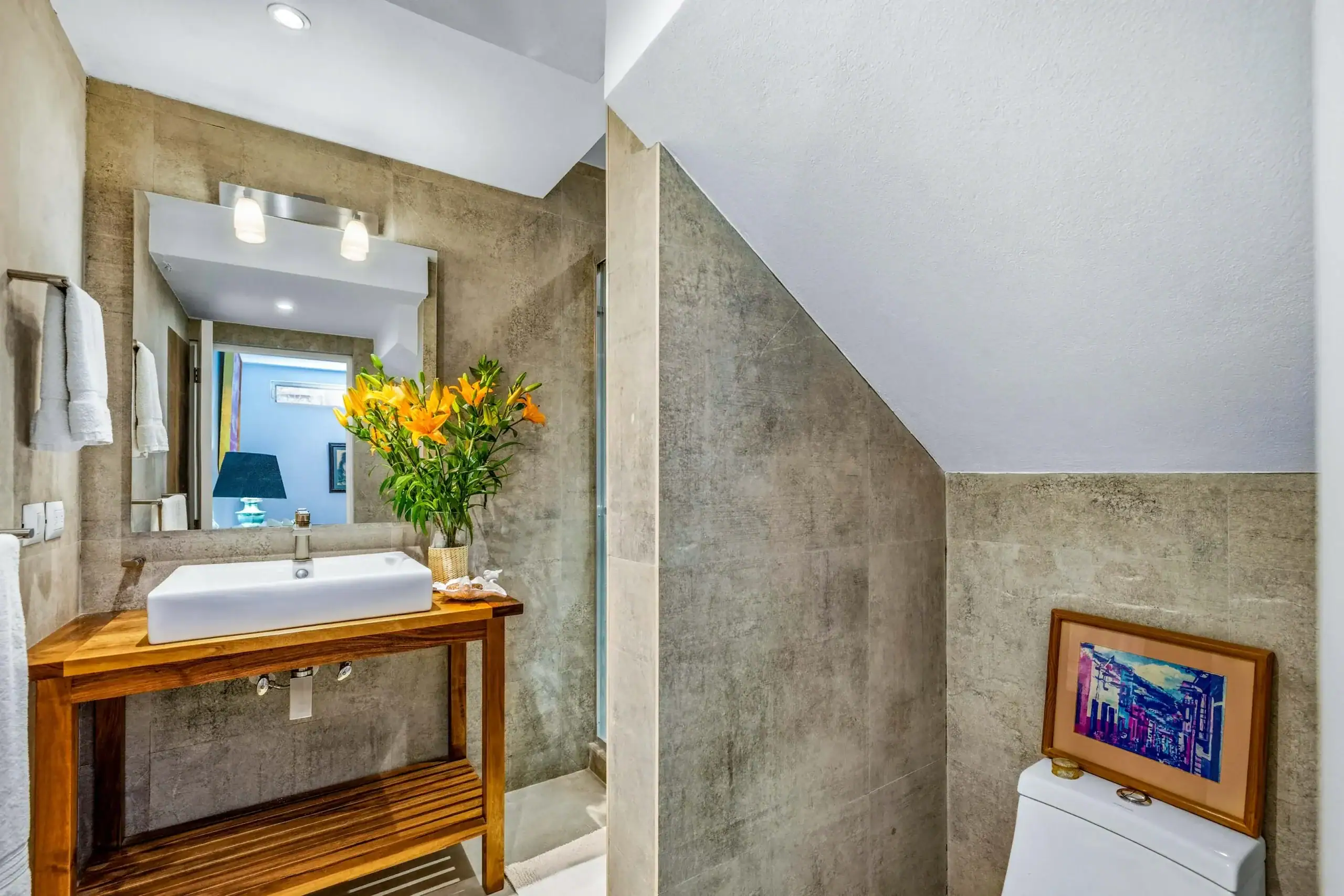 Modern bathroom with a wood-framed sink console, a white rectangular sink, and a vase of orange lilies on the counter, reflected in a large mirror with towels on a rack nearby, San Miguel de Allende