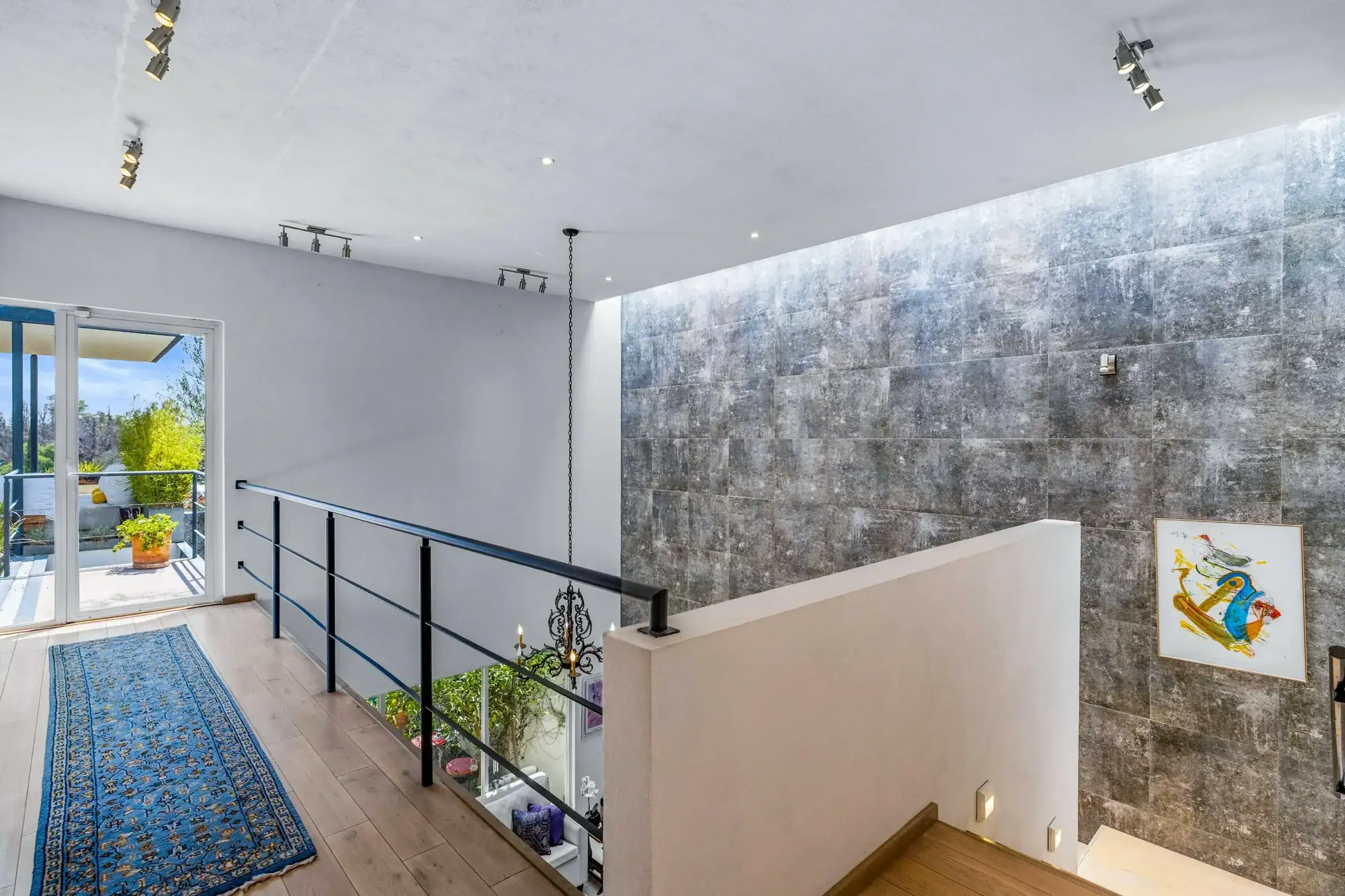 Modern stairwell landing with black metal railing, gray stone accent wall, hanging chandelier, track lighting, and a blue patterned runner rug leading to glass doors opening to a sunny patio with potted plants, San Miguel de Allende