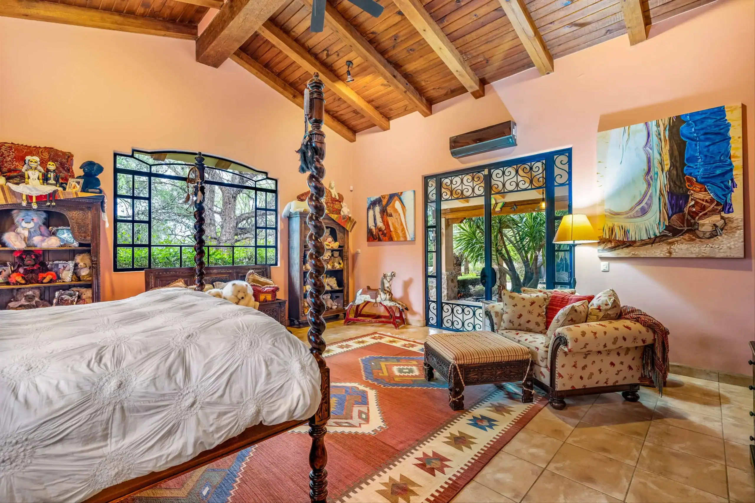 Bedroom with a carved four-poster bed, pink walls, and exposed wooden beam ceiling, plus a colorful rug and seating area, San Miguel de Allende