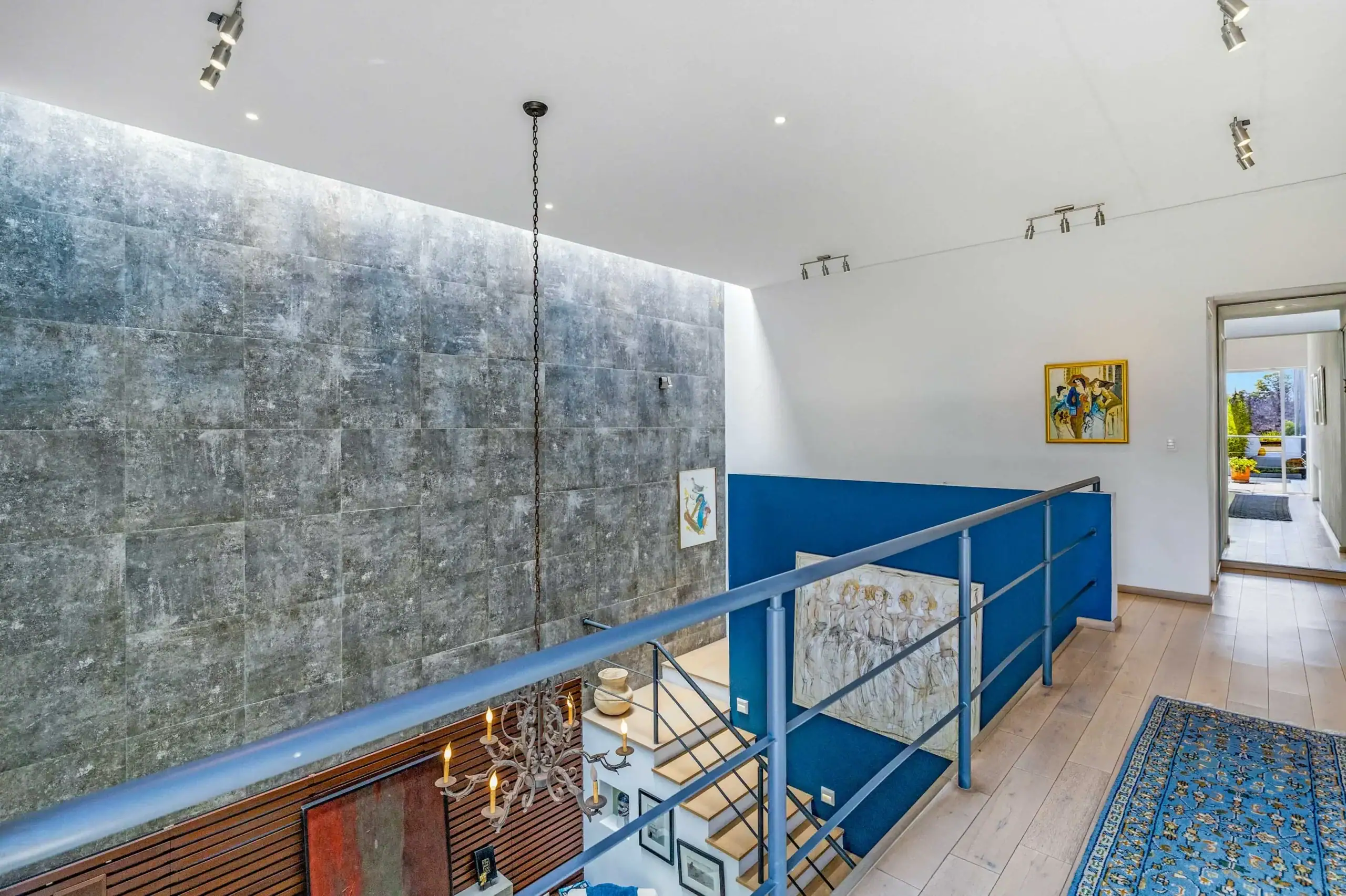 Two-story interior stairwell with blue railing, gray tiled wall, and hanging wrought-iron chandelier; doorway to an outdoor area visible at end, San Miguel de Allende