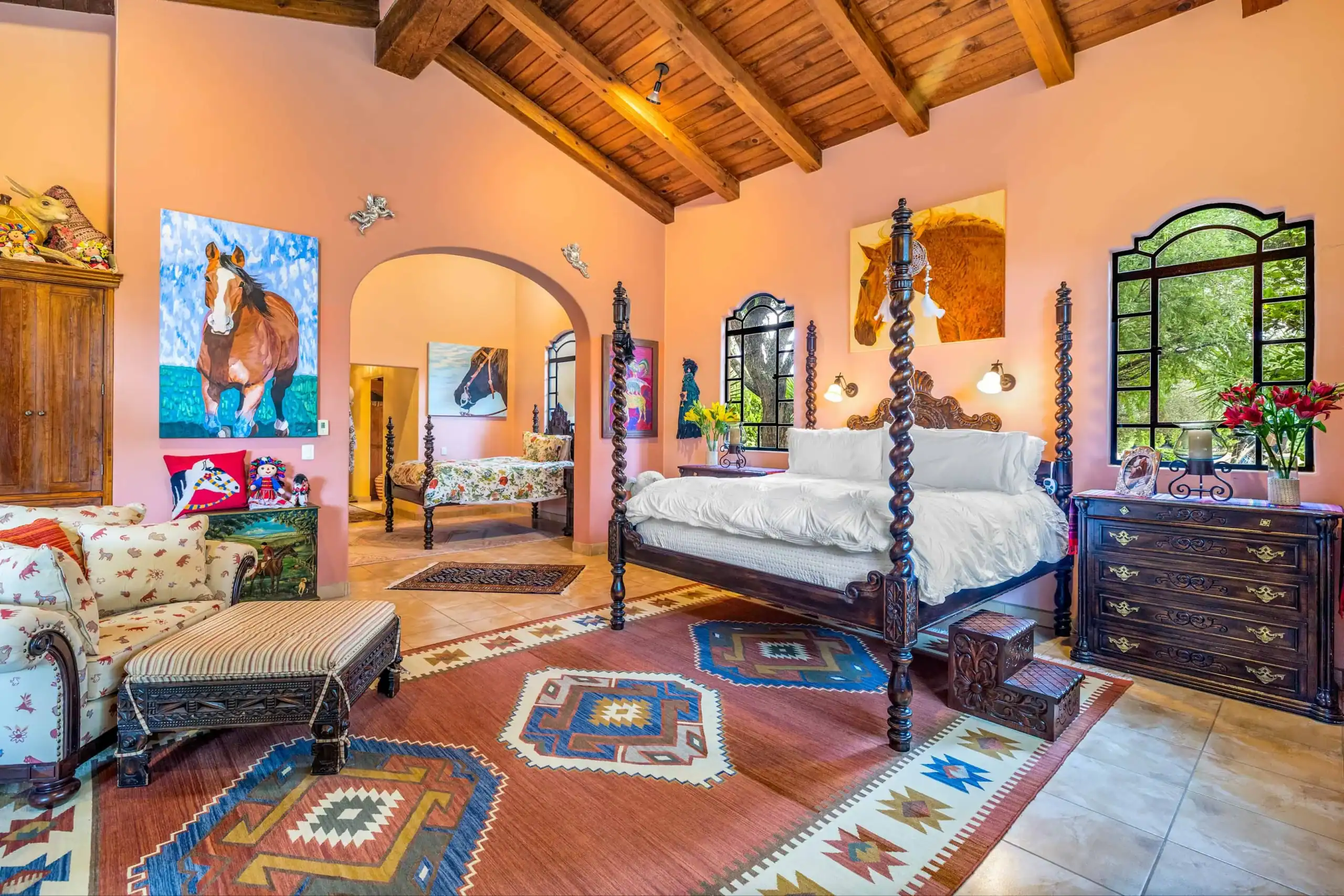 Rancho Lusitanos, bedroom with pink walls, a dark carved wood four-poster bed, a patterned kilim rug, horse paintings, an arched doorway and a wood beam ceiling, San Miguel de Allende