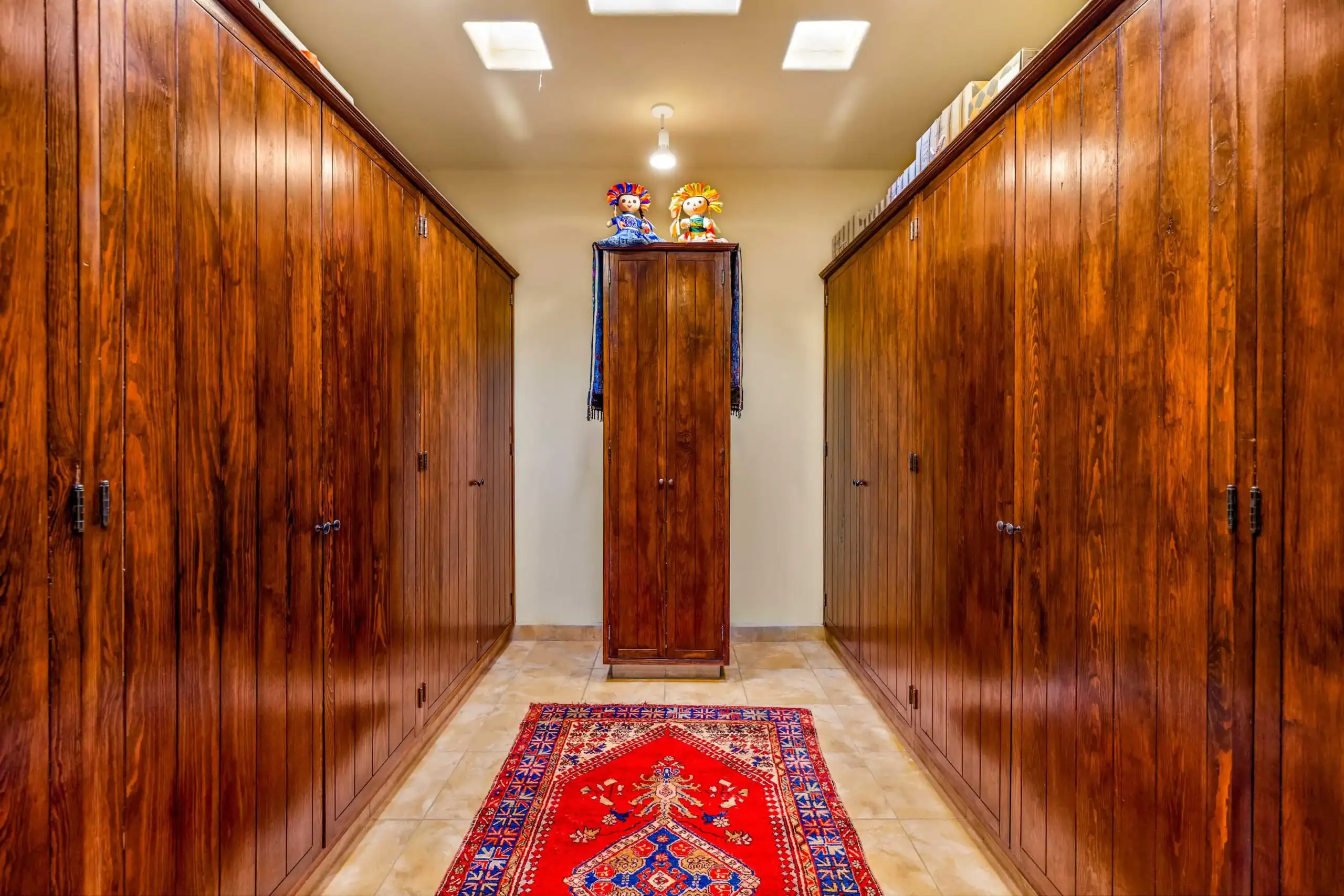 Narrow hallway flanked by tall wooden closets; a central cabinet holds two colorful dolls, with a red patterned rug on the tiled floor, San Miguel de Allende