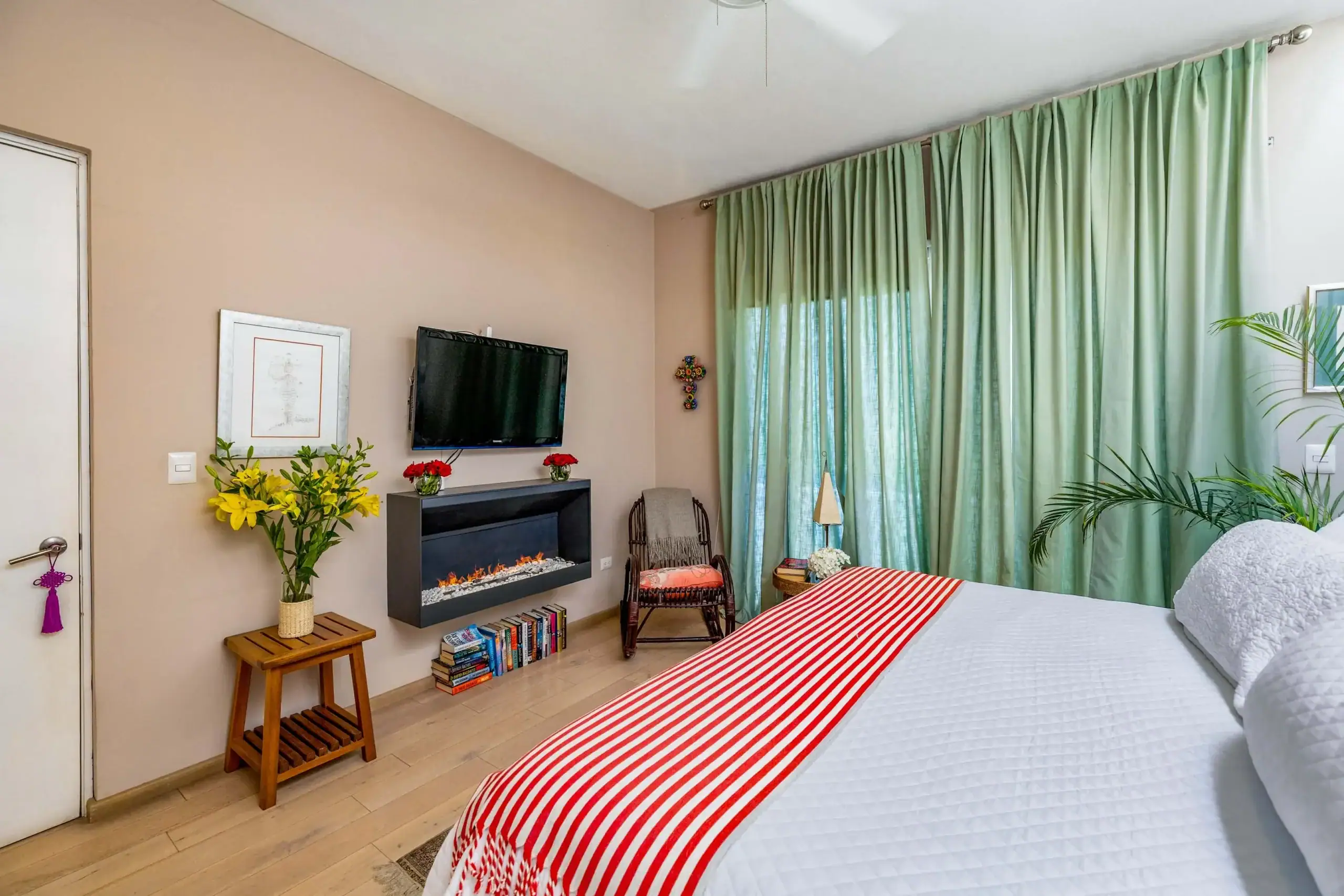 Bedroom corner with a wall-mounted TV above a rectangular electric fireplace, pink walls, and large green curtains; potted plants and flowers on the mantel add color, San Miguel de Allende