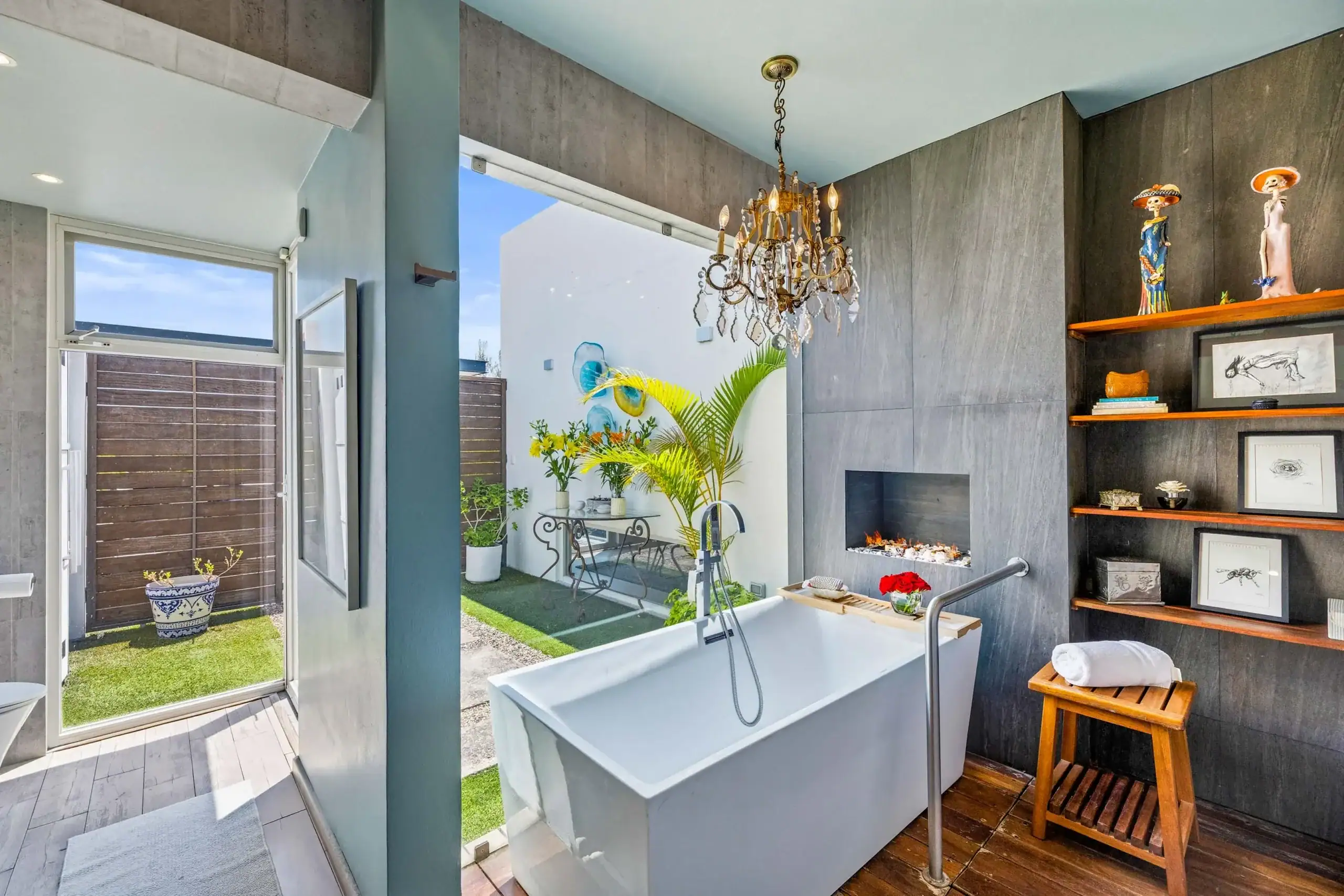 Modern bathroom interior with a white tub, dark gray walls, and a brass chandelier, open to a sunny courtyard with plants outside, San Miguel de Allende